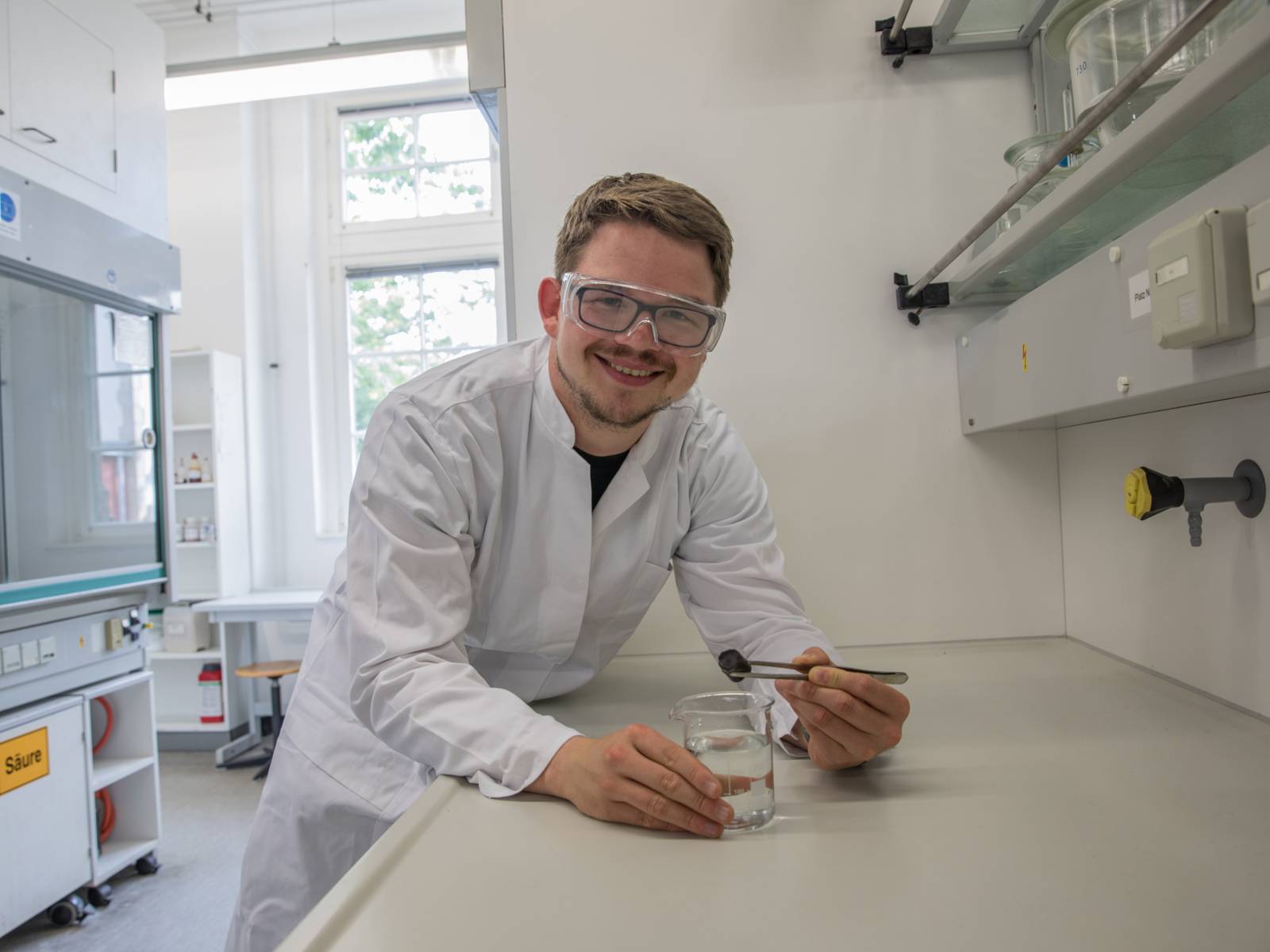 Mann mit Schutzbrille und weißem Kittel in einem Labor.