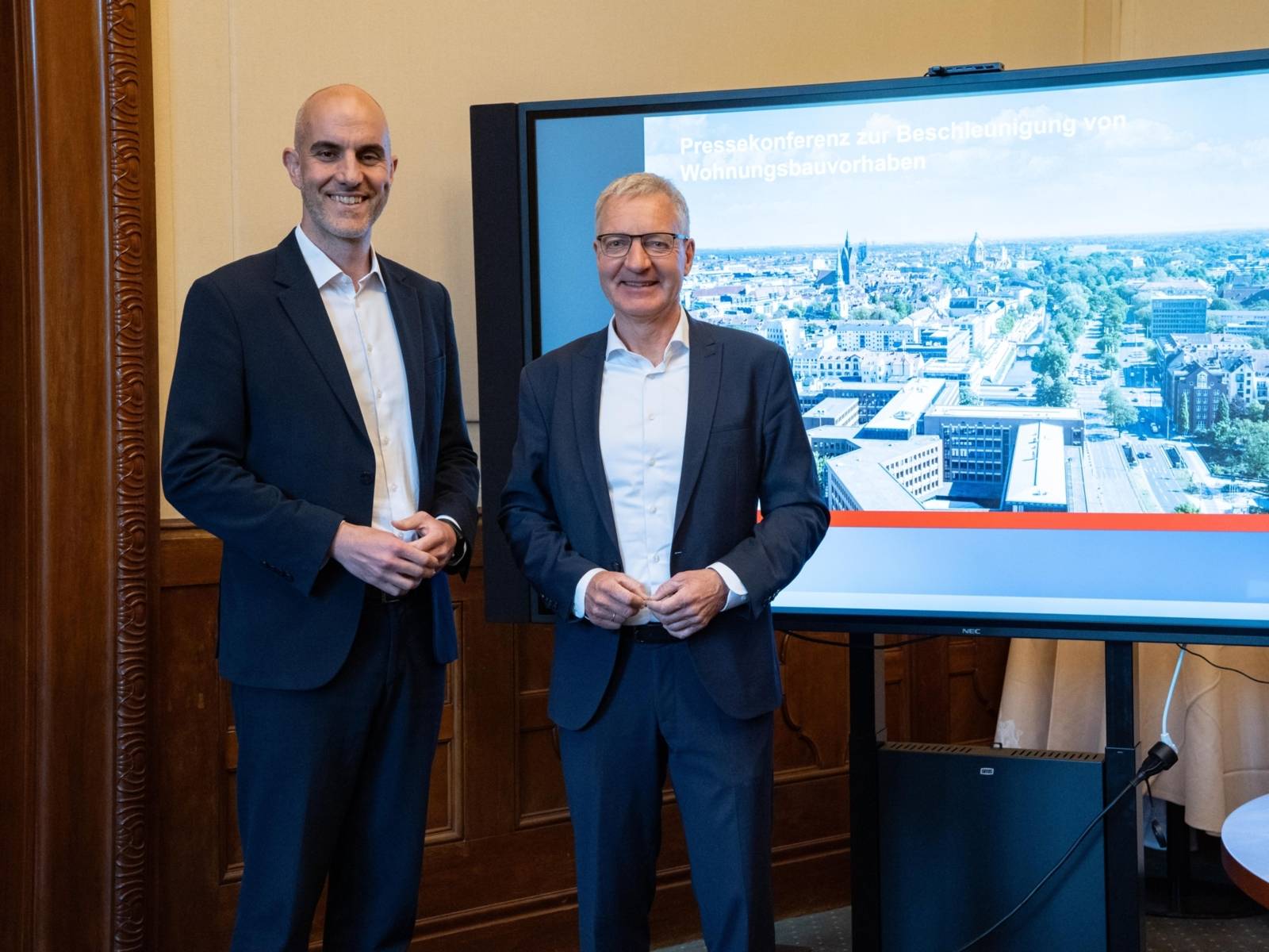 Zwei M&auml;nner in Anz&uuml;gen stehen in einem Raum neben einem gro&szlig;en Monitor mit einem Bild auf eine Stadt aus der Vogelperspektive, dar&uuml;ber steht Pressekonferenz zur Beschleunigung von Wohnungsbauvorhaben.