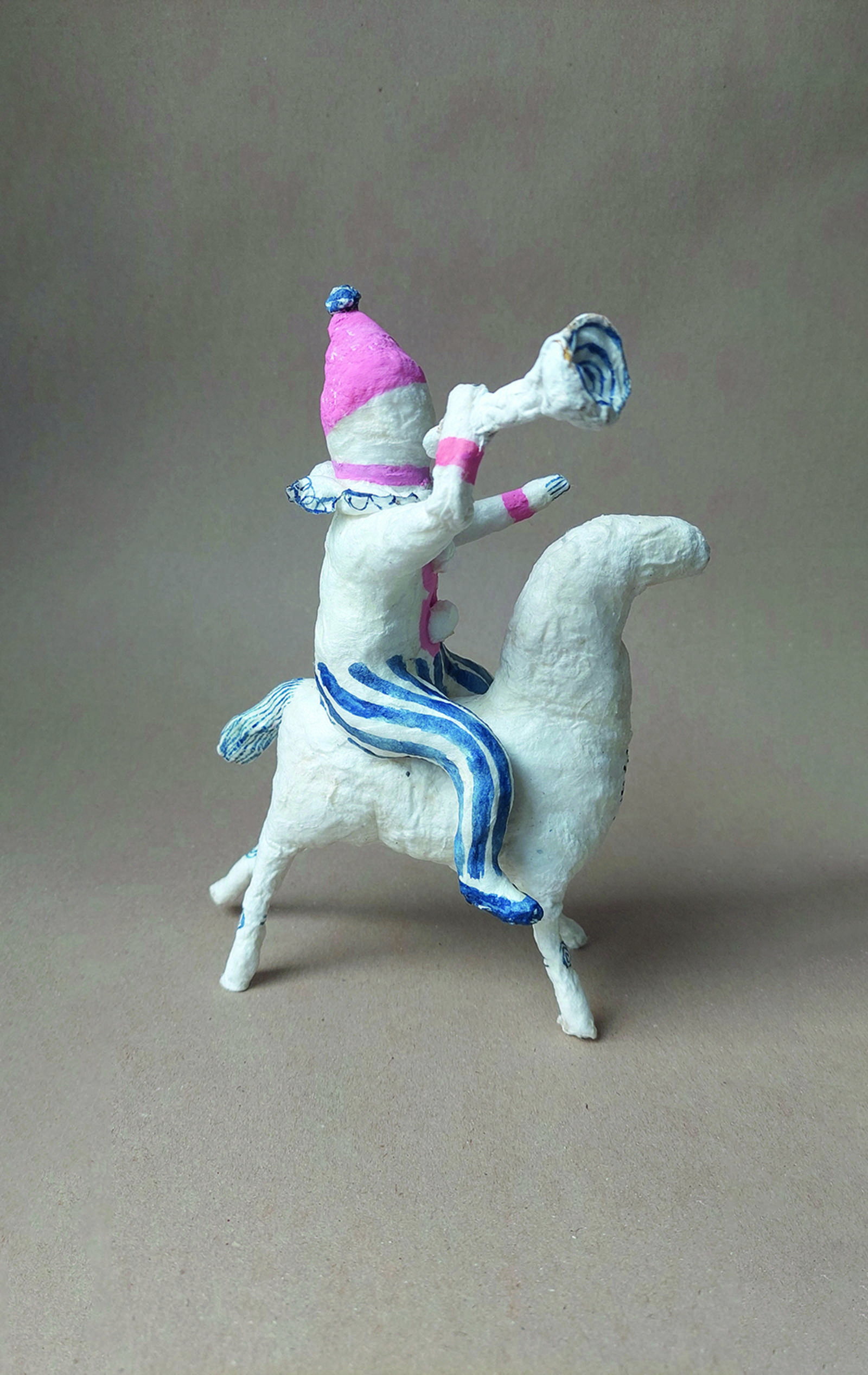Zu sehen ist eine kleine Figur gefertigt von Kira Kotliar. Auf einem weißen vierbeinigem Tier sitzt ein kleines Männchen und bläst in eine Tröte.