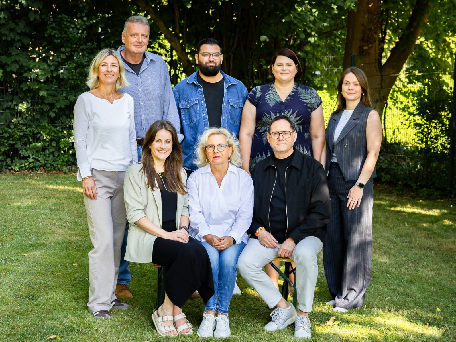 Acht Personen stehen auf einer Rasenfläche oder sitzen auf einer Bank vor den Stehenden.
