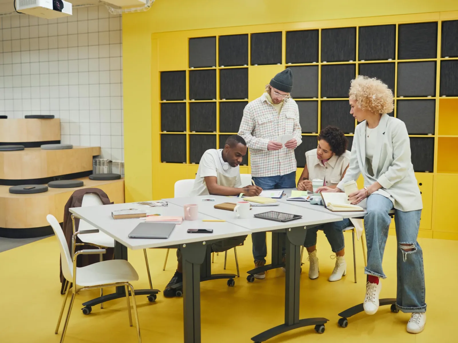 Vier Personen gruppieren sich um einen Gruppentisch. Anscheinend sind es zwei Frauen und zwei Männer. Sie tragen helle Kleidung. Sie konzentrieren sich auf Arbeitsunterlagen auf dem Tisch. Die Personen haben unterschiedliche Hautfarben. Der Raum, in dem sie sich befinden, ist in hellem gelb gehalten.