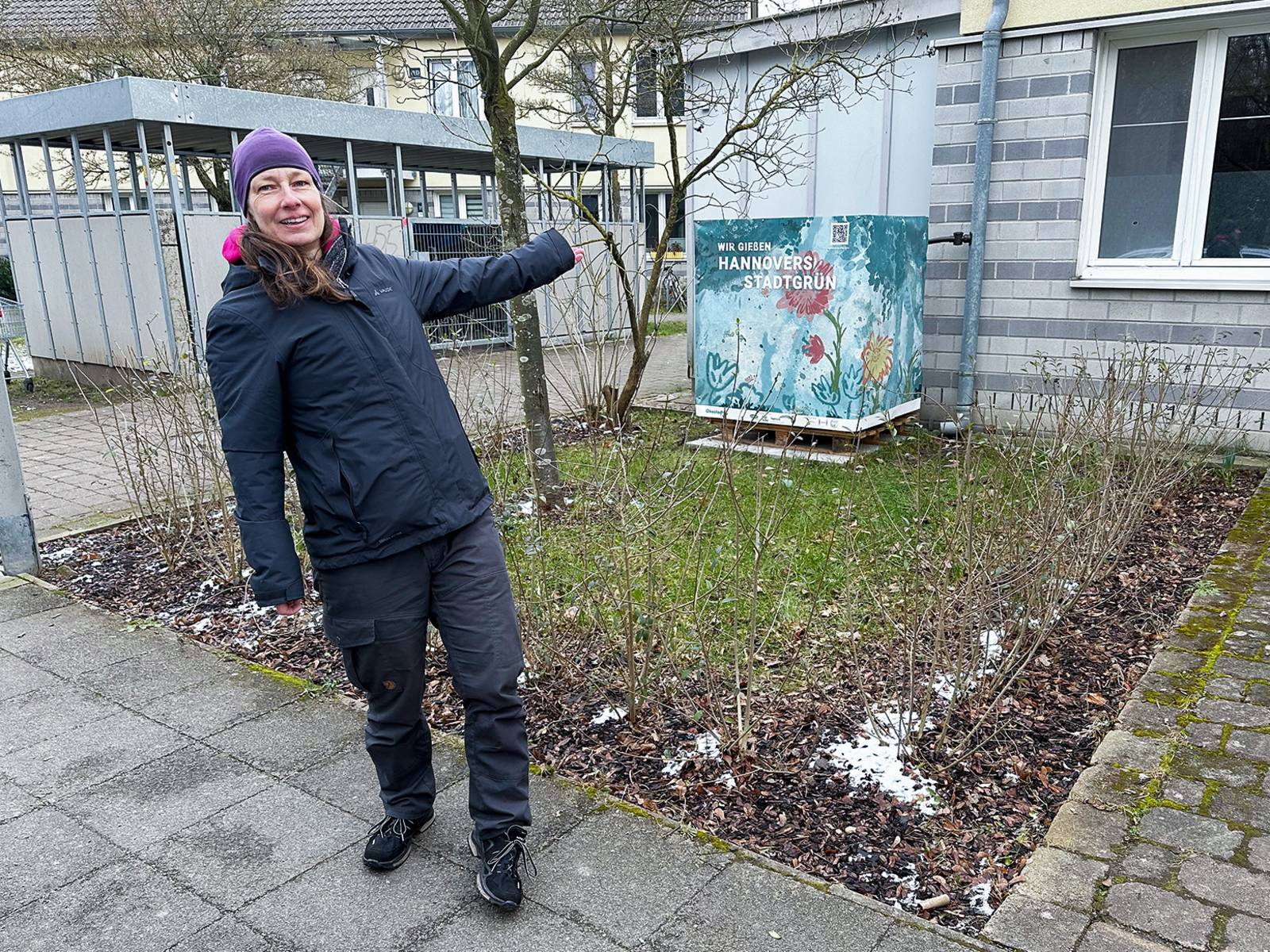 Eine  Frau, die auf einen bedruckten Kanister mit Wasser zeigt. Darauf steht "Wir gießen Hannovers Stadtgrün", außerdem ein QR-Code.