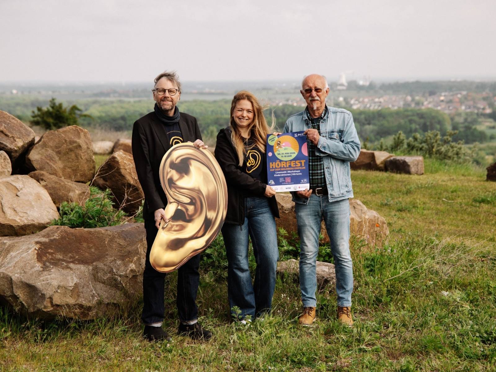 Zwei Männer und eine Frau stehen auf einer Wiese und halten ein Plakat und ein goldenes Ohr-Plakat in die Kamera. Im Hintergrund ist die Stadt und der Horizont zu sehen.