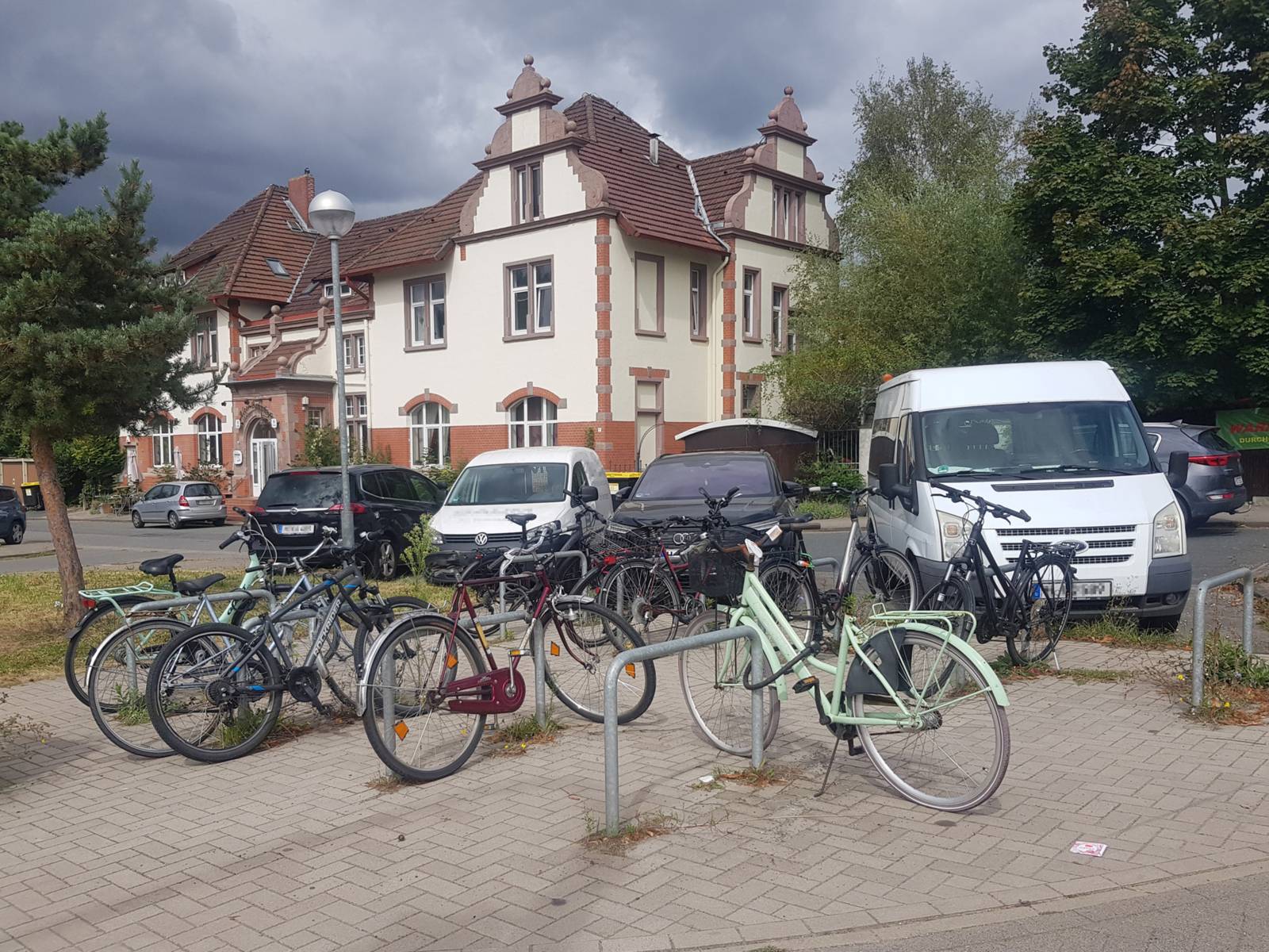 Auf einem Platz mit neun Fahrradb&uuml;geln stegen mehrere angeschlossene R&auml;der, dahinter eine Stra&szlig;e mit parkenden Autos und ein Wohnhaus, rechts und links B&auml;ume.