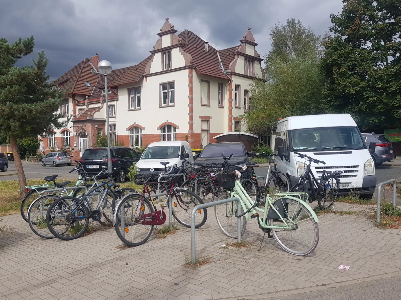 Auf einem Platz mit neun Fahrradb&uuml;geln stegen mehrere angeschlossene R&auml;der, dahinter eine Stra&szlig;e mit parkenden Autos und ein Wohnhaus, rechts und links B&auml;ume.