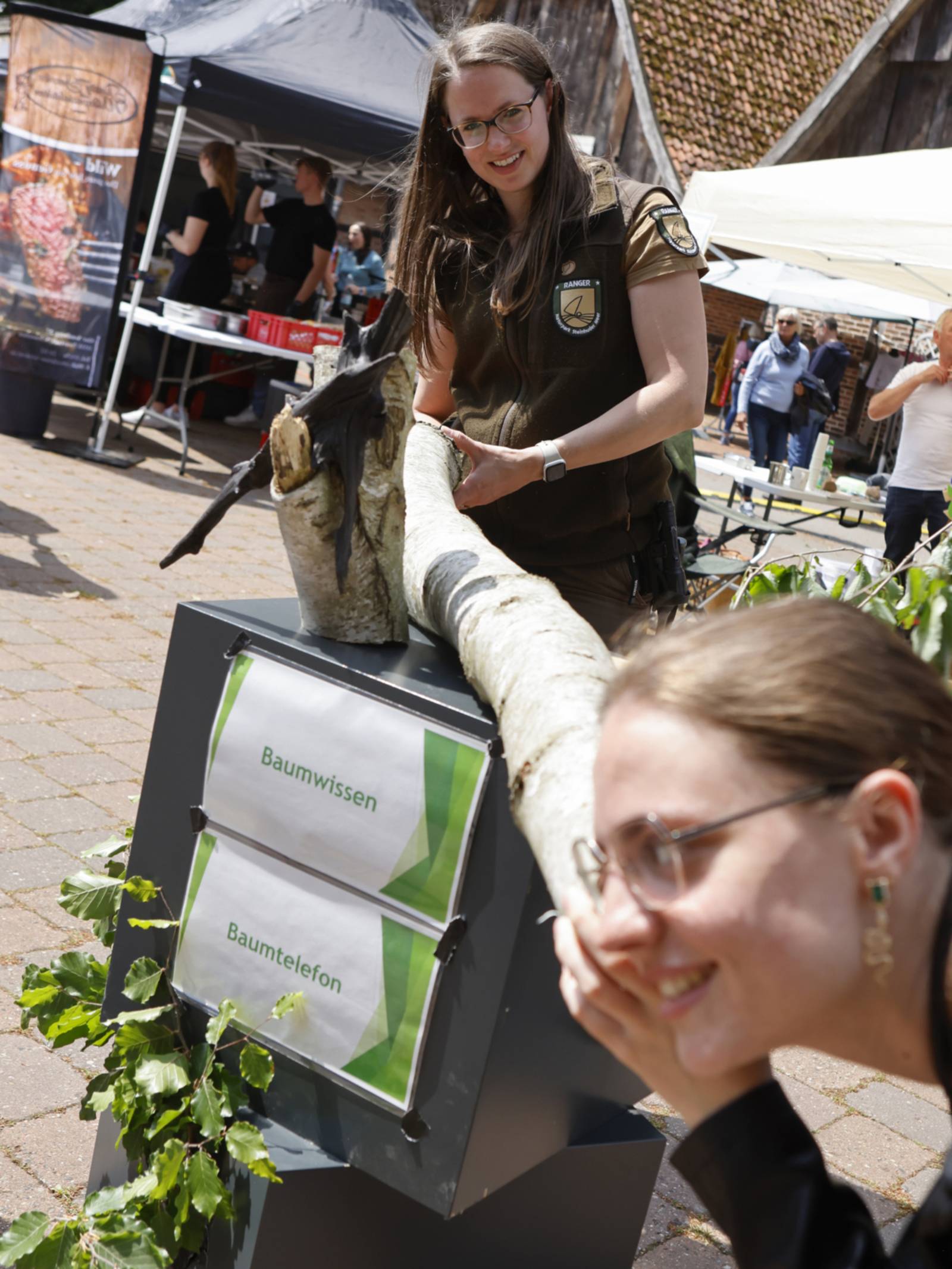 Eine Person h&auml;lt einen Baumstamm, eine weitere Person legt das Ohr an den Baumstamm.