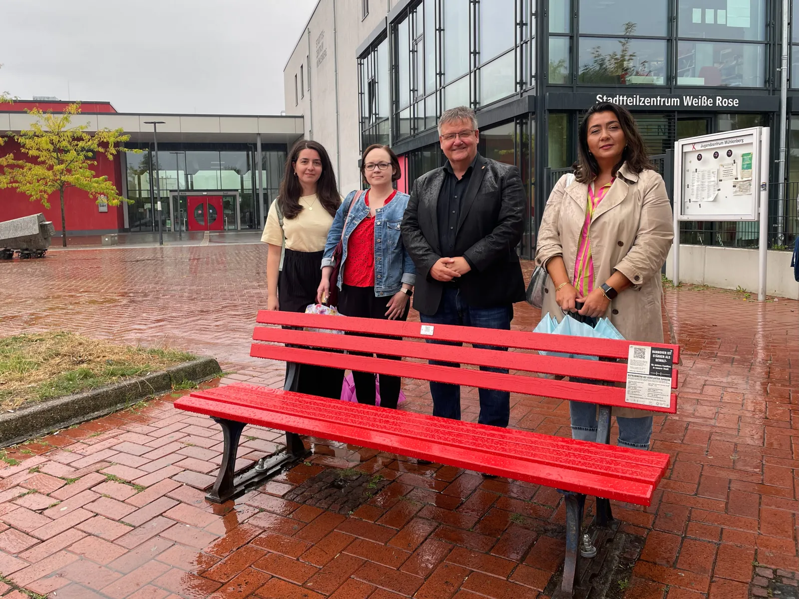 Vier Frauen und ein Mann stehen hinter einer rot angestrichenen Sitzbank auf einem Platz.