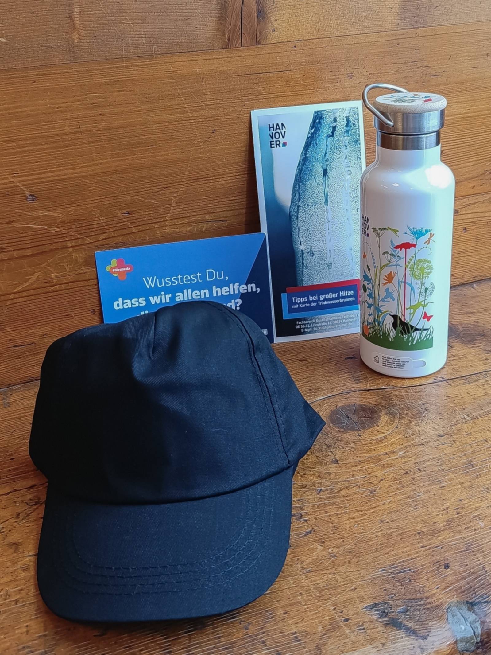 Eine Schirmm&uuml;tze, eine Trinkflasche aus Edelstahl ein Flyer und eine Postkarte zusammengestellt auf einer Holzunterlage