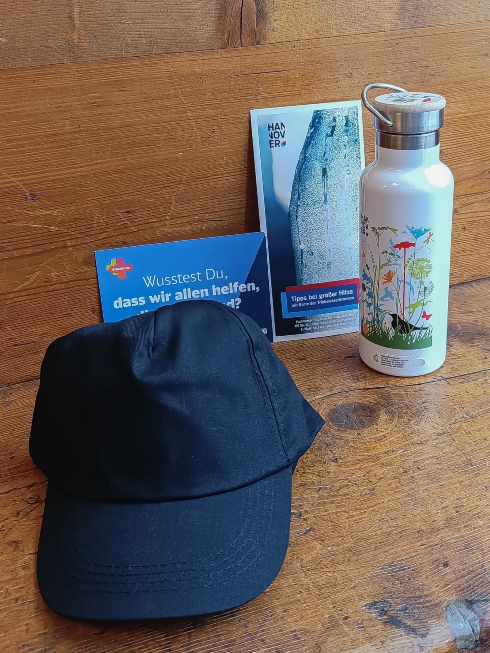 Eine Schirmm&uuml;tze, eine Trinkflasche aus Edelstahl ein Flyer und eine Postkarte zusammengestellt auf einer Holzunterlage
