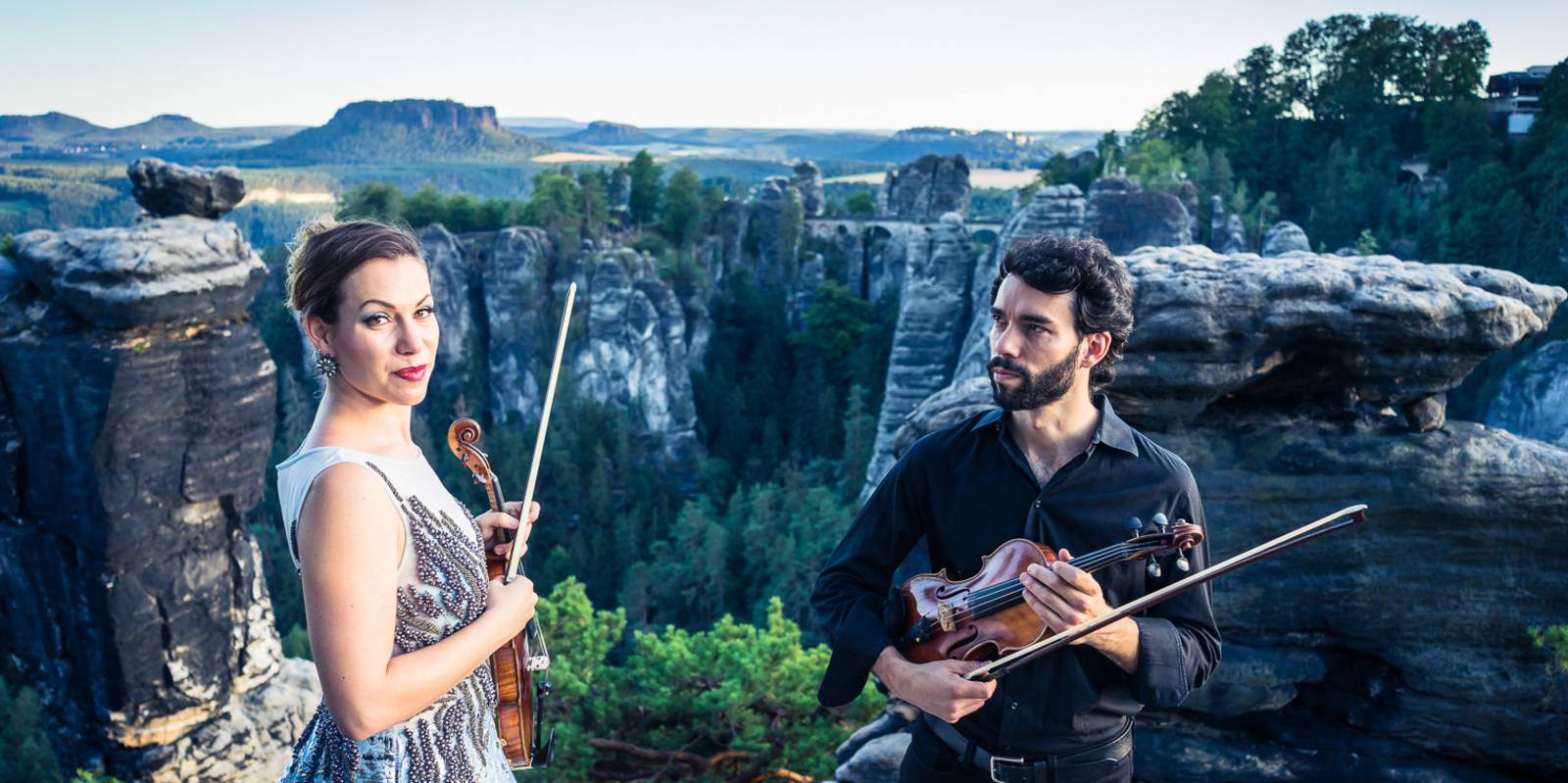 Eine Musikerin und ein Musiker halten jeweils eine Violine und einen Bogen in der Hand, hinter ihnen ist eine Landschaft mit grünen Bäumen, Felsen und einem Abgrund.