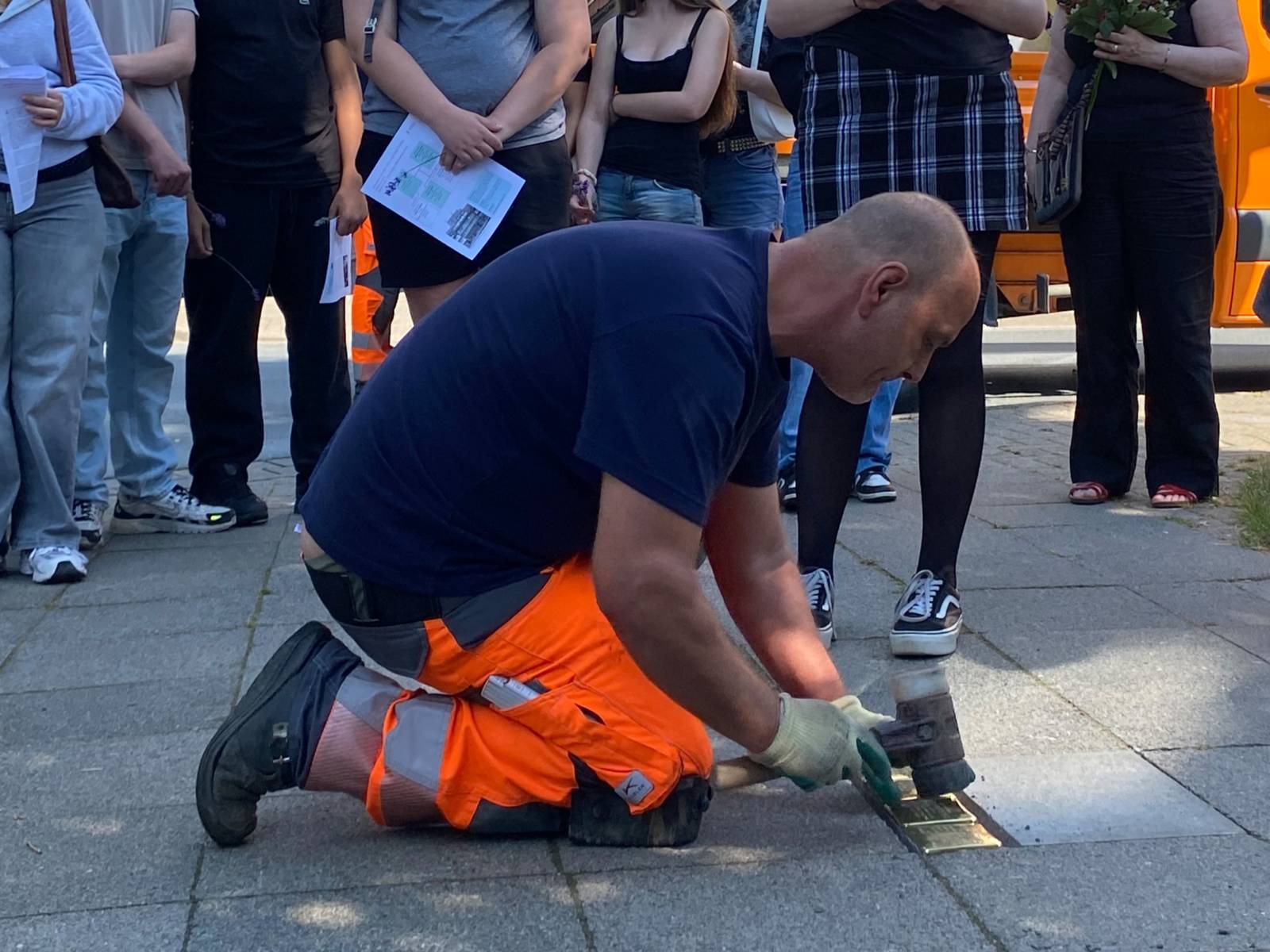 Ein Mann in blauem Shirt und oranger Hose kniet auf dem Fu&szlig;weg und schl&auml;gt mit einem Gummihammer goldene quadratische Steine in den Boden. Dahinter sind die Beine einer Gruppe von jungen Menschen zu erkennen.