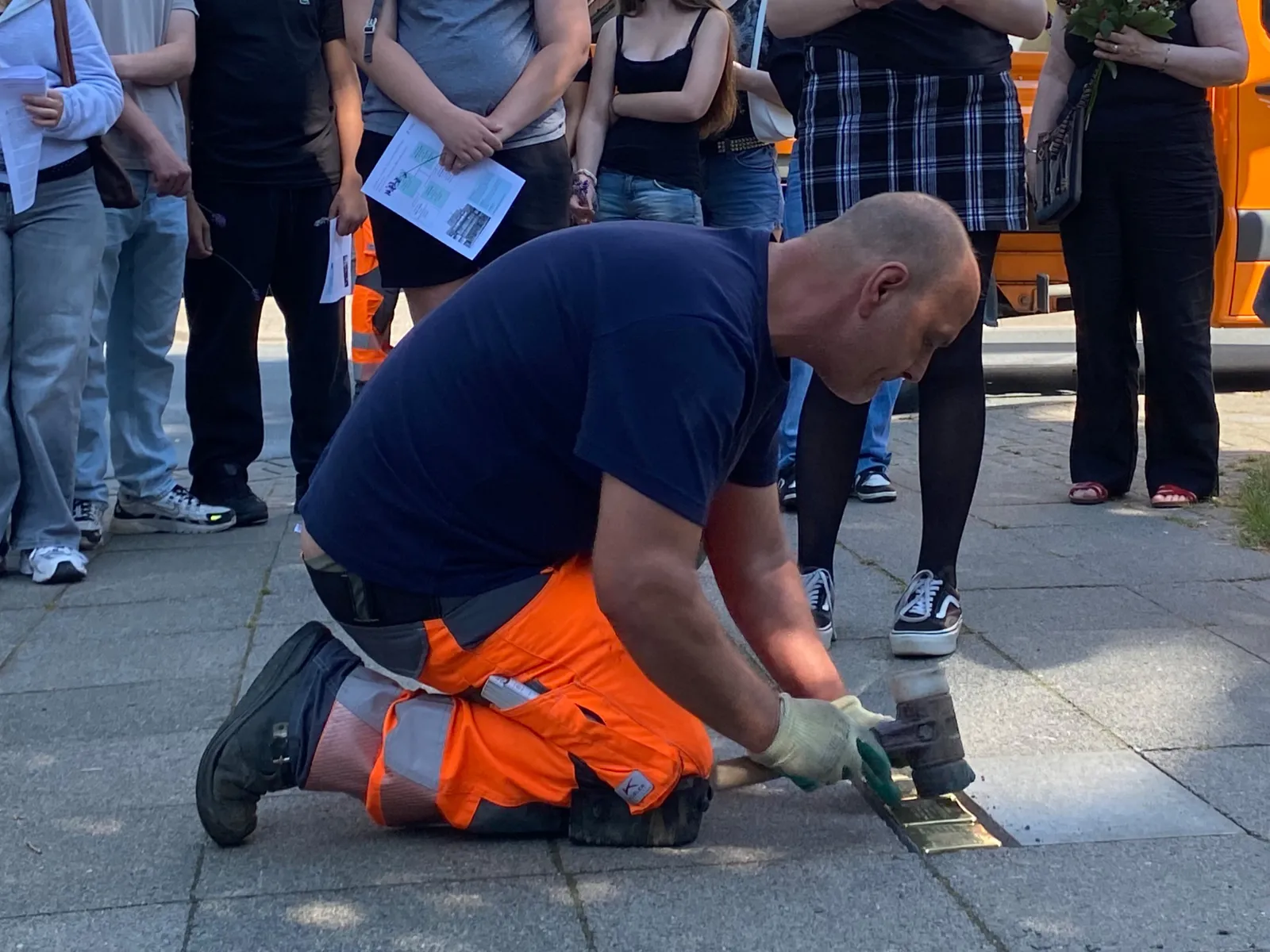 Ein Mann in blauem Shirt und oranger Hose kniet auf dem Fu&szlig;weg und schl&auml;gt mit einem Gummihammer goldene quadratische Steine in den Boden. Dahinter sind die Beine einer Gruppe von jungen Menschen zu erkennen.