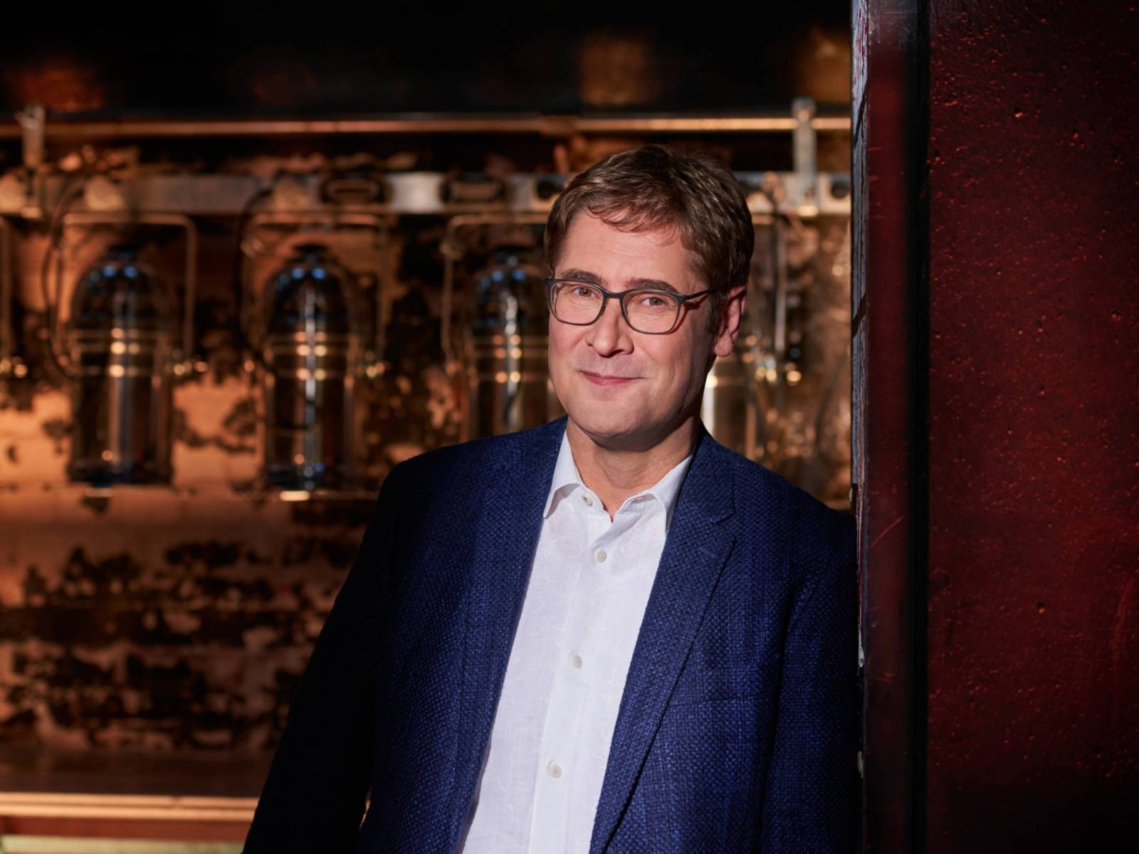 Eine Person mit Brille, die einen blauen Anzug trägt, lehnt an einer Wand.