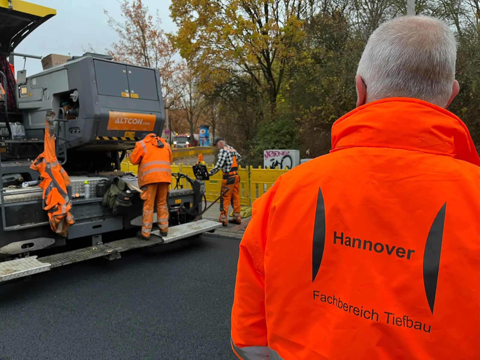 Ein Mann in einer leutend orangen Jacke ist von hinten zu sehen, auf der Jacke steht Hannover, Fachbereich Tiefbau. Er blickt auf einen breiten fahrenden Wagen, auf dem ein Mann steht, daneben steht ein Mann mit einer Schaufel. Unten der Bodenbelag ist tief schwarz glänzend.