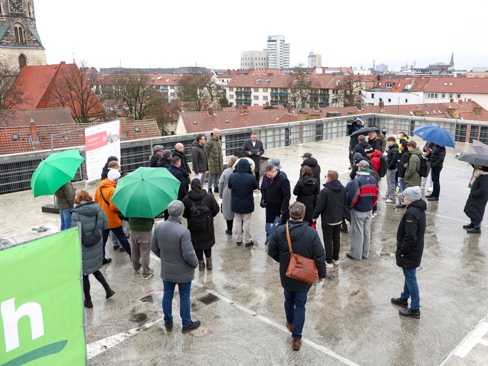 Auf einem Dach stehen Menschen, zum Teil mit Schirmen. Ihr Blick ist f&uuml;nf Personen gerichtet, von denen ein Mann spricht. Im Hintergrund sind D&auml;cher und ein Kirchturm zu sehen.