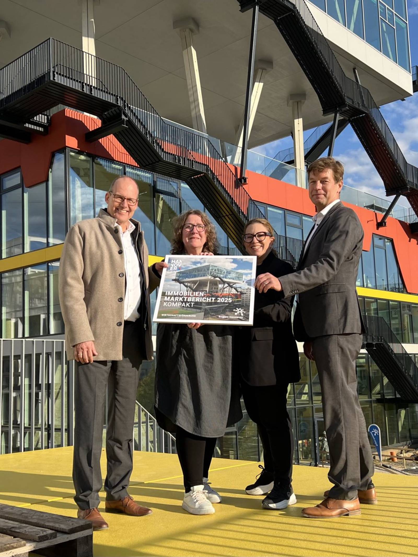 Vier Personen stehen vor einem Geb&auml;ude und halten das Deckblatt des Immobilienmarktberichtes 2025 der Region Hannover.