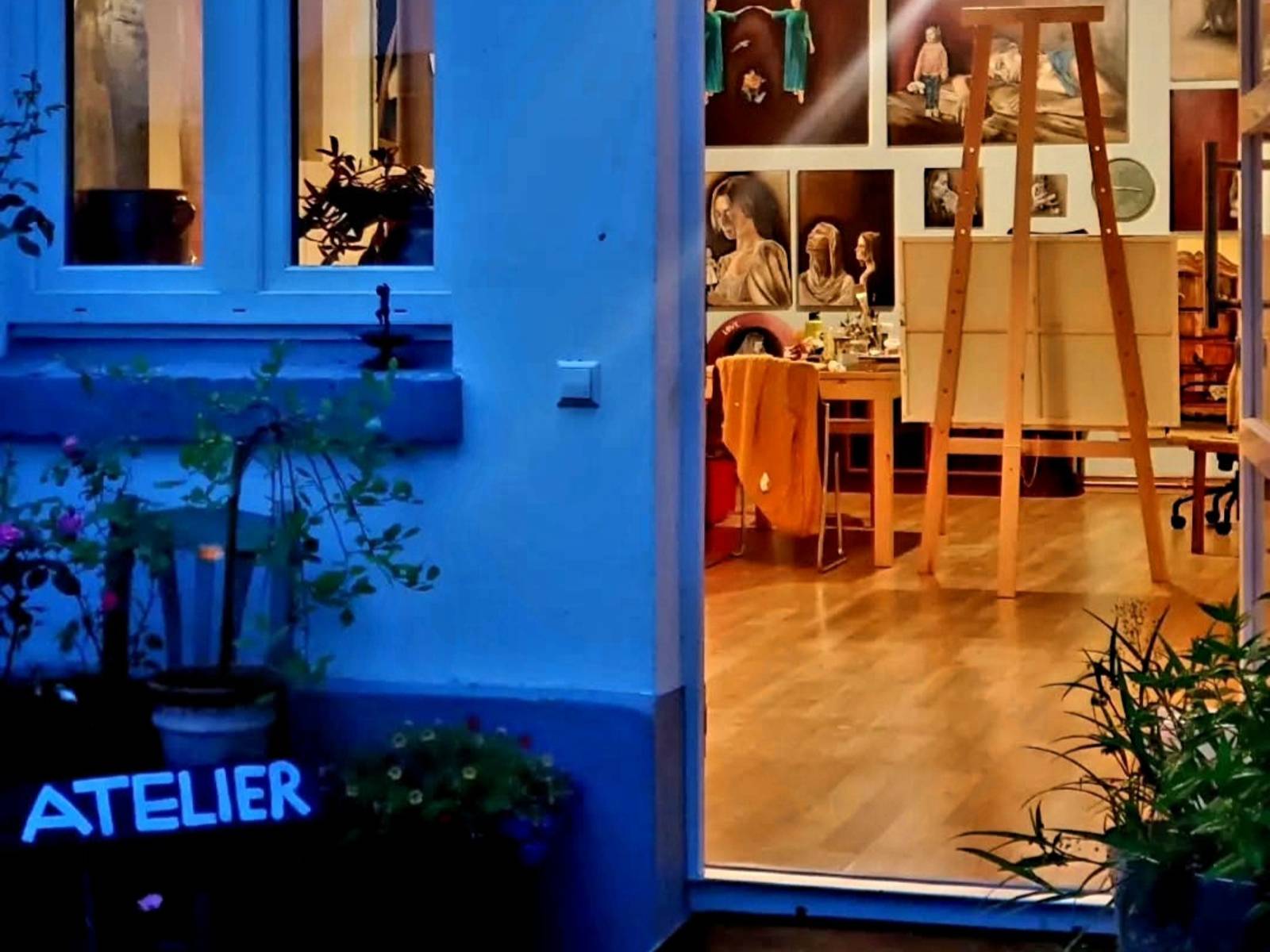 Blick durch eine ge&ouml;ffnete T&uuml;r in das Atelier eines K&uuml;nstlers: Werke h&auml;ngen an den W&auml;nden, Arbeitsmaterialien wie Pinsel und Staffeleien sind im Raum verteilt und es gibt M&ouml;bel wie ein Sofa und einen Schrank mit Malutensilien.