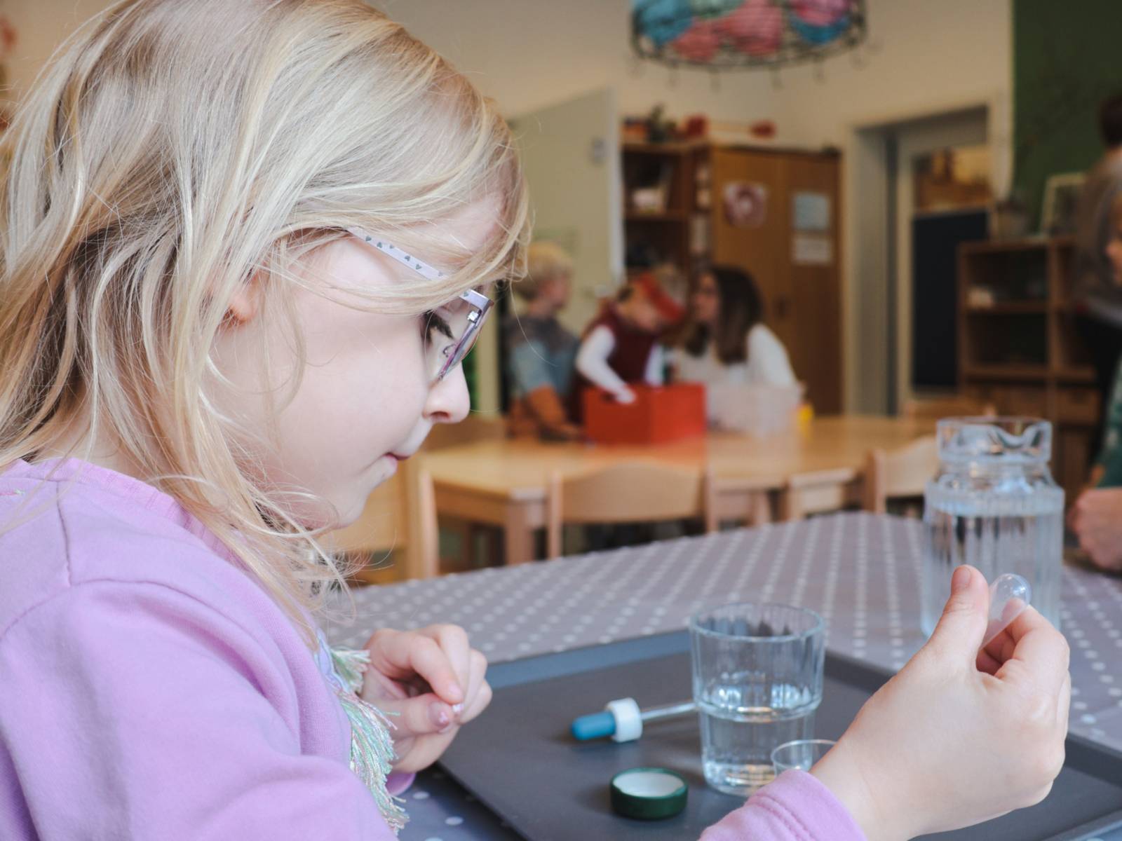 Ein Mädchen sitzt an einem Tisch, in der Hand hält das Mädchen eine Pipette, auf dem Tisch sind Gläser mit Wasser.