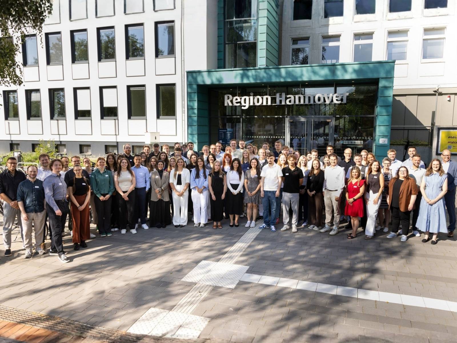 78 Nachwuchskräfte und andere Mitarbeitende der Region Hannover stehen vor dem Eingang des Regionshauses und schauen in die Kamera, die etwas oben fotografiert.