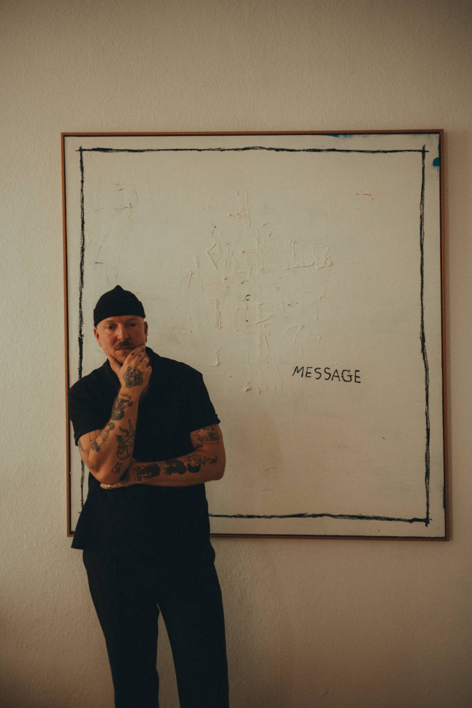 Zu sehen ist der K&uuml;nstler Timo Hinkelmann. Er steht vor seinem gro&szlig;en Gem&auml;lde, einer wei&szlig; gestrichene Leinwand mit den Worten &bdquo;Message&ldquo;.