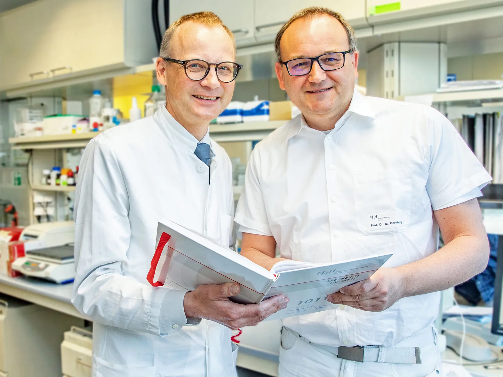 Zwei Männer in weißen Kitteln in einem Labor halten gemeinsam ein Buch in den Händen.