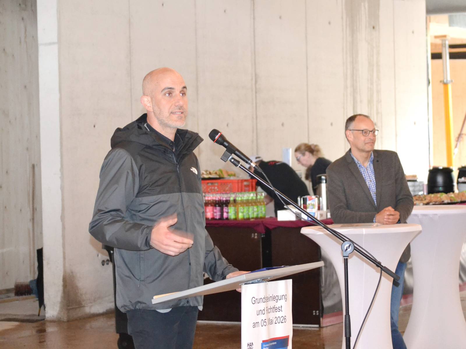 In einem Raum mit einer kahlen Betonwand steht ein Mann vor einem Rednerpult und spricht. Rechts daneben steht ein weiterer Mann auf einen Stehtisch gelehnt. Hinter ihn ein Stand mit Flaschen udn zwei Personen in schwarzen Hemdblusen.