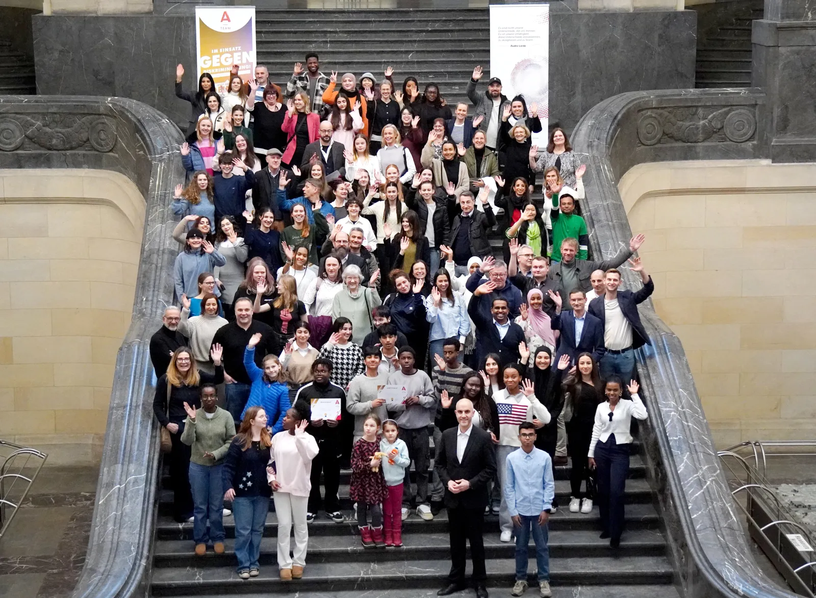 Eine große Gruppe von Menschen steht auf einer barocken Treppe und winkt in die Kamera.