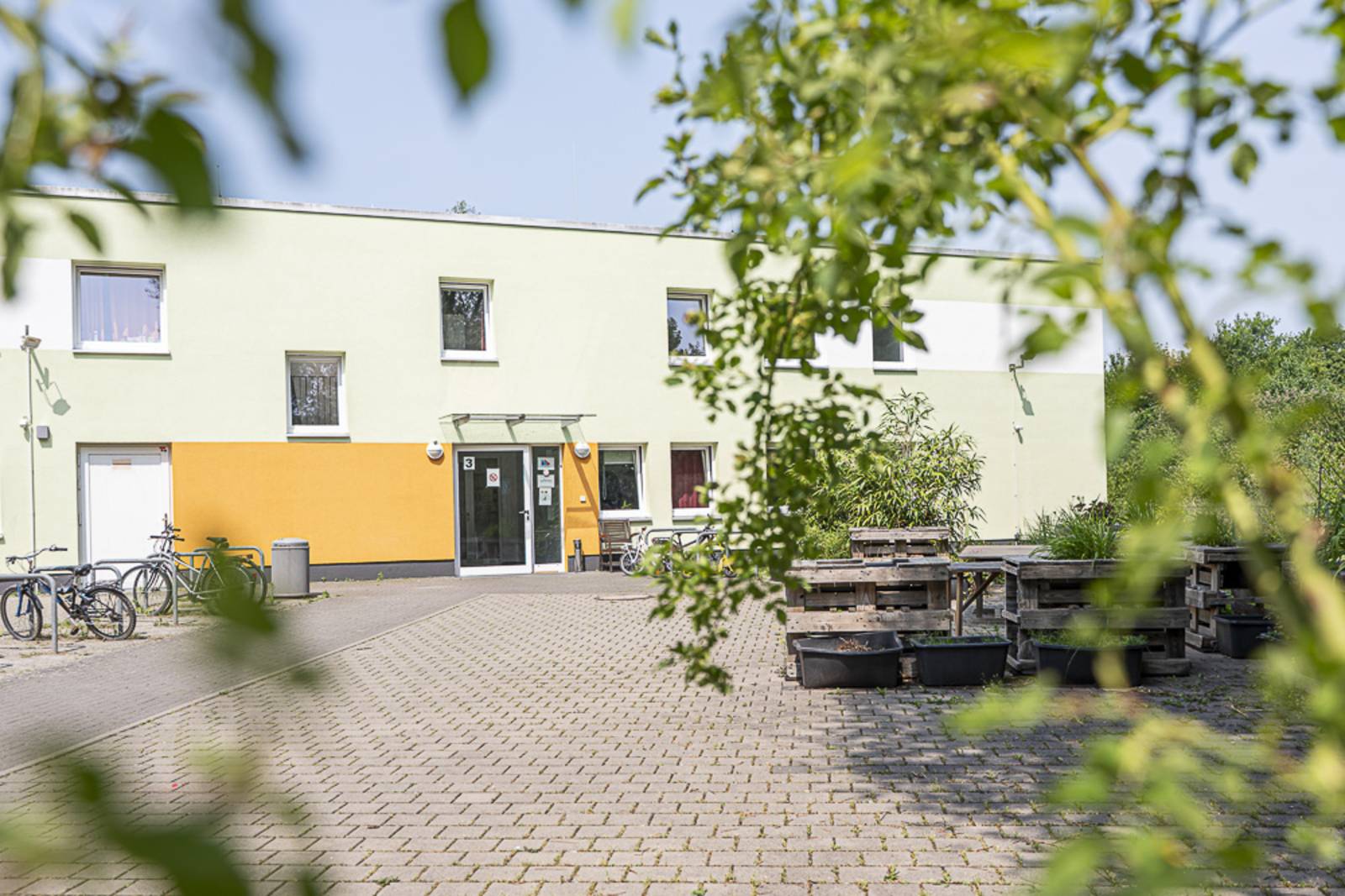 Ein flaches, zweistöckiges Gebäude mit einigen Fenstern und einem überdachten Eingang. Das Gebäude ist mintgrün und hell-orange gestrichen. Vor dem Gebäude einige Hochbeete, Bänke und Tische.