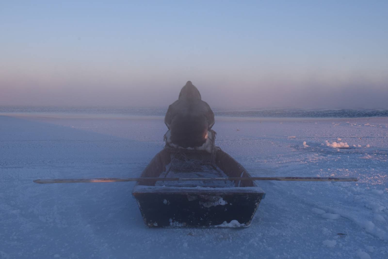Zu sehen ist eine Fotografie einer Eislandschaft bei Sonnenuntergang. Auf einem Boot sitzt eine Person in dicker Winterkleidung mit dem Rücken zum Betrachter.