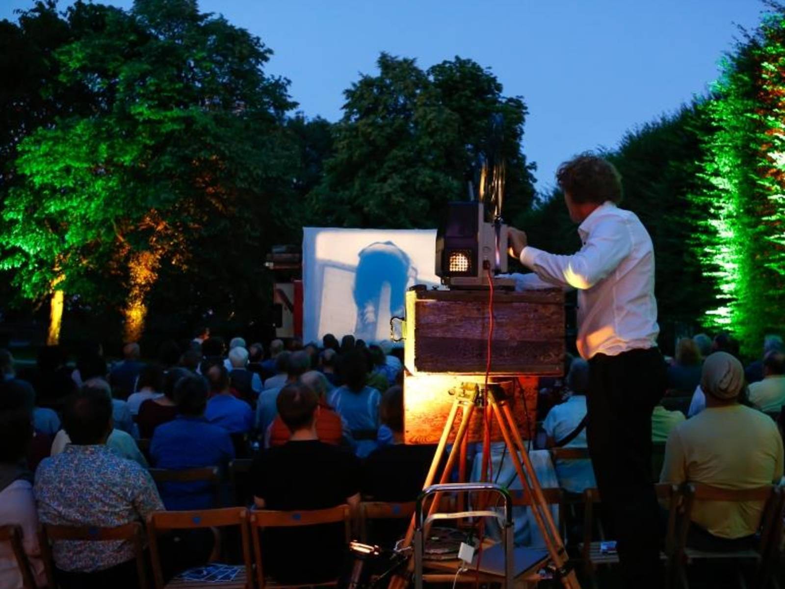 Ein Mann bedient einen Projektor uns wirft die Szene eines Films unter freiem Himmel auf eine Leinwand, Publikum verfolgt den Film.