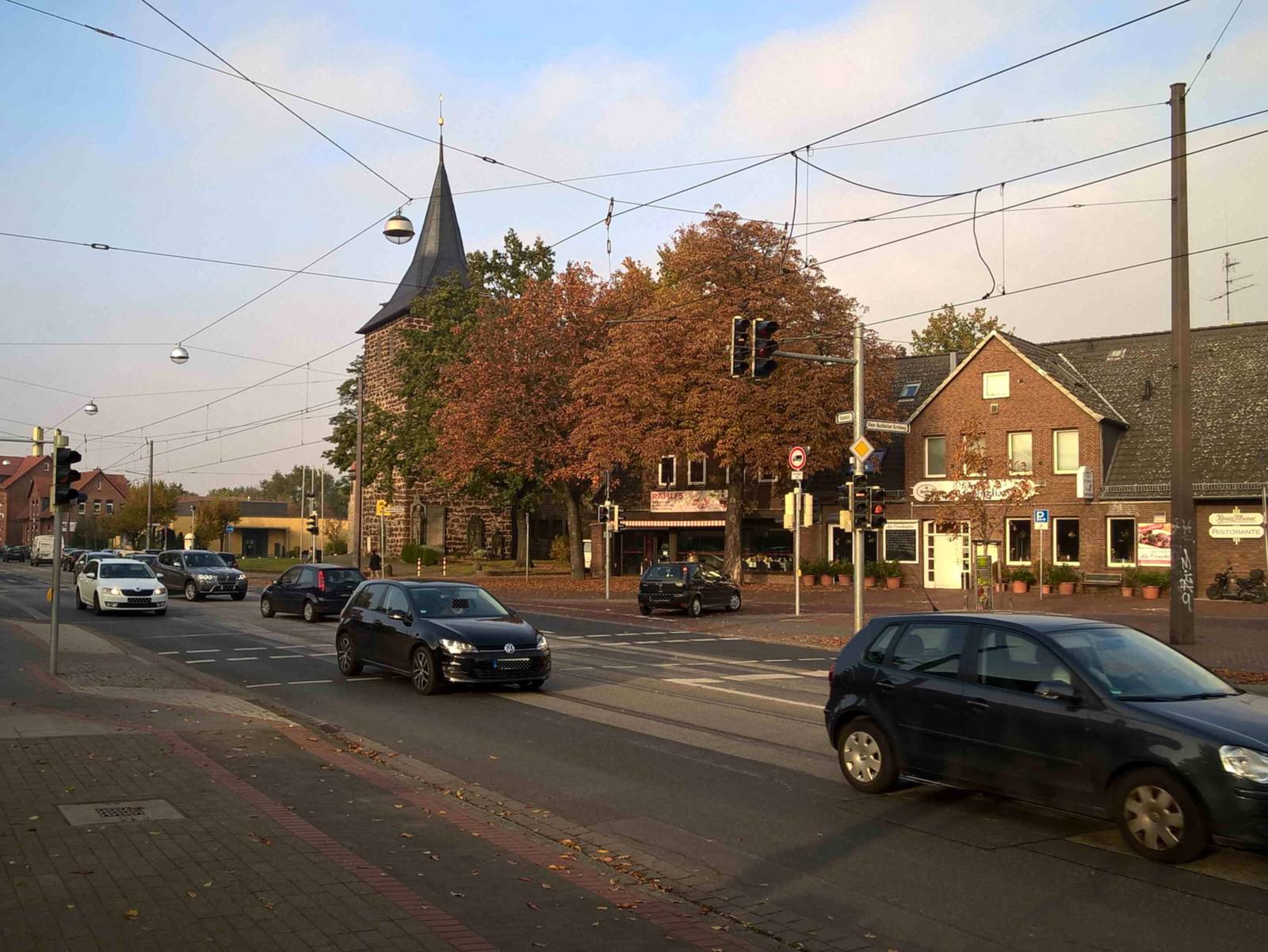 Auf dem Bild ist eine Kreuzung in Bothfeld zu sehen. Auf der Straße befinden sich Autos. Im Hintergrund ist ein altes Gebäude mit einem großen alten Baum zu sehen. Die Blätter des Baums sind herbstlich. Neben dem Haus ist eine Kirche zu sehen.
