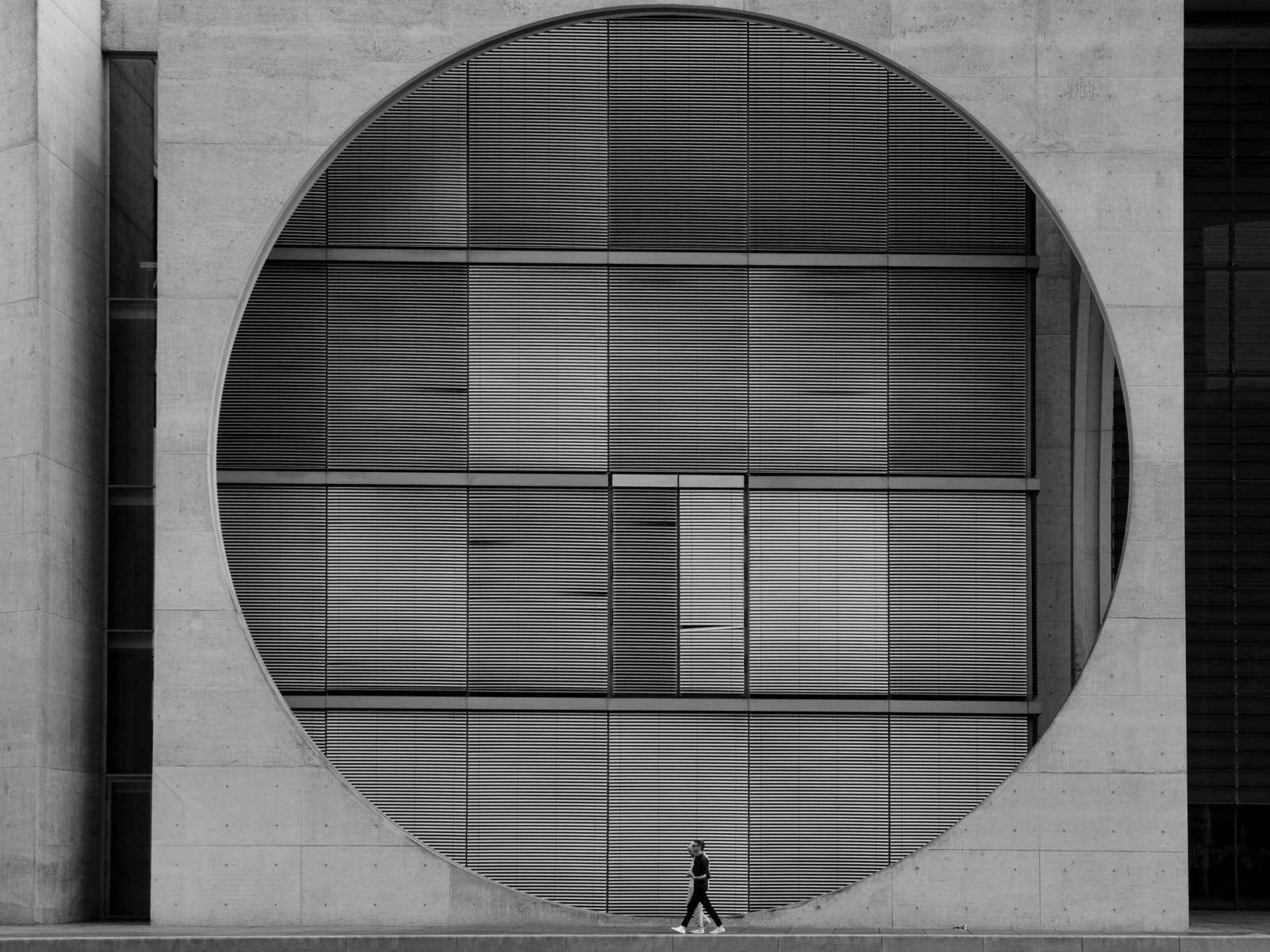 Zu sehen ist eine Schwarz-Weiß-Fotografie von Diana Schmidt: vor einem großen urbanen Gebäude sind zwei Menschen am unteren Rand zu sehen, die Spazieren gehen.