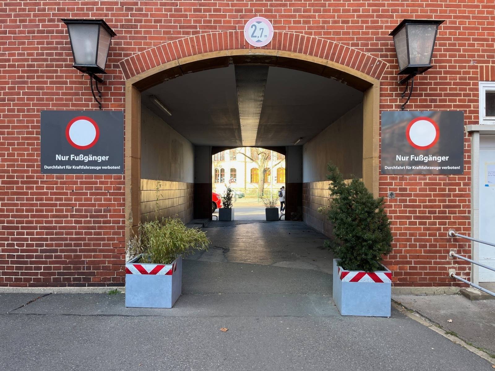Ein Torbogen mit einem Durchgang in einem roten Klinkergeb&auml;ude. Rechts und links des Bogens stehen Pflanztr&ouml;ge, dar&uuml;ber sind am Haus Schilder "Nur Fu&szlig;g&auml;nger" angebracht. Hinter dem Durchgang ist Fu&szlig;weg und der Ausschnitt eines weiteren Geb&auml;udes zu sehen.