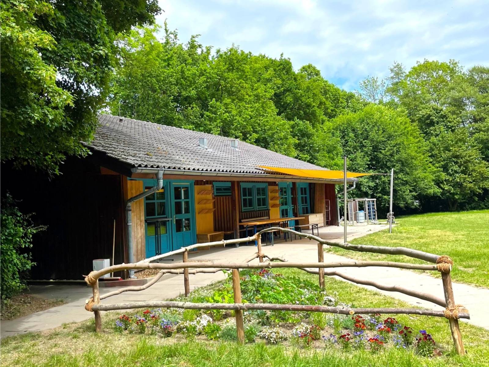 Eine Holzhütte mit blauen Fenster- und Türelementen. Dahinter liegen Bäume und davor eine Wiese mit einem umzäunten Blühbeet.