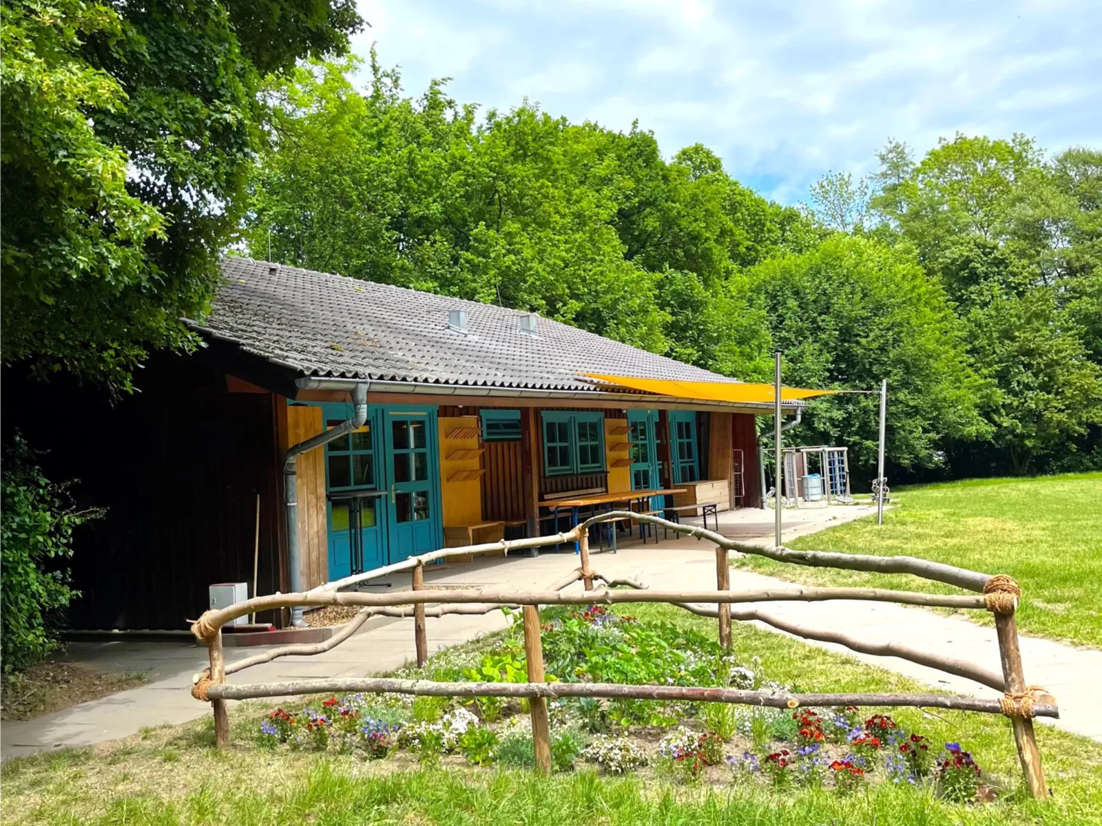 Eine Holzhütte mit blauen Fenster- und Türelementen. Dahinter liegen Bäume und davor eine Wiese mit einem umzäunten Blühbeet.