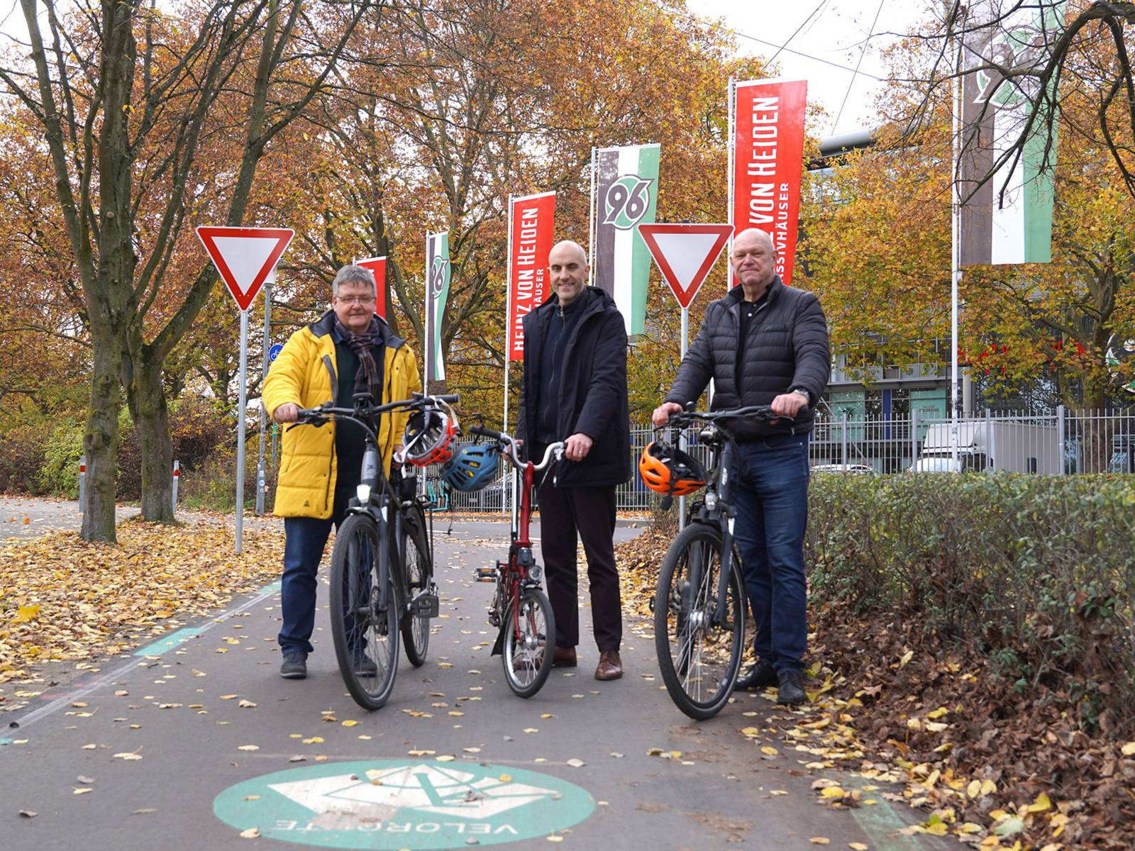 Drei Männer stehen neben ihren Fahrrädern auf einem Radweg, auf dem vor ihnen ein grüner Kreis mit dem Wort Veloroute steht. Um sie sind Büsche und Bäume mit Herbstlaub, hinter ihnen befinden sich Fahnenmasten mit Fahnen mit den Aufschriften Heinz von Heiden und 96.