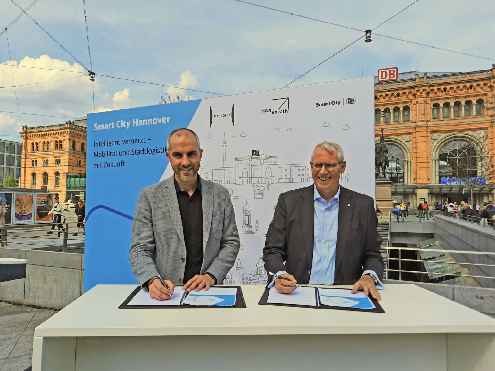 Hannovers OB Belit Onay und Vorstandsvorsitzender von DB Station&Service, Bernd Koch, unterzeichnen an einem Stand vor dem Hauptbahnhof ein Dokument.