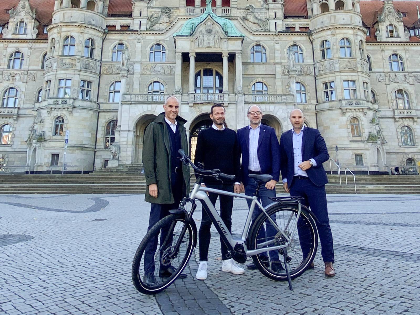 Vier Männer stehen vor dem Neuen Rathaus in Hannover, im Vordergrund ein silbernes E-Bike.