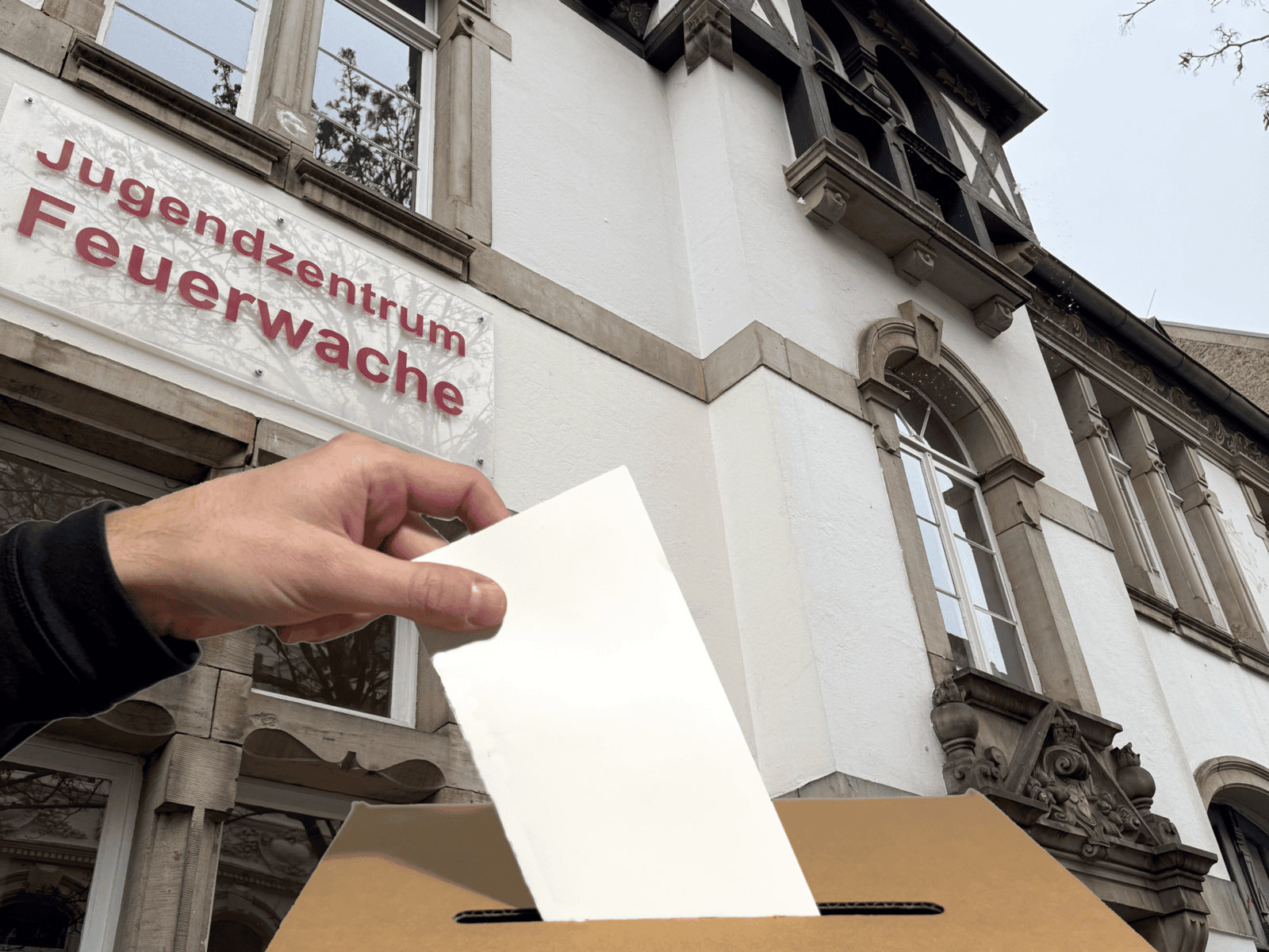 Eine Hand wirft einen Umschlag in eine Wahlurne. Im Hintergrund ein Jugendzentrum "Feuerwache".
