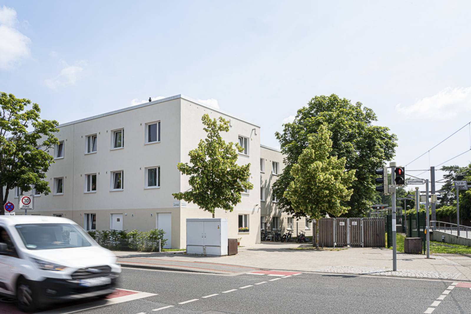 Ein beiges, dreistöckiges Gebäude mit Flachdach. Davor einige Bäume und eine befahrene Straße.