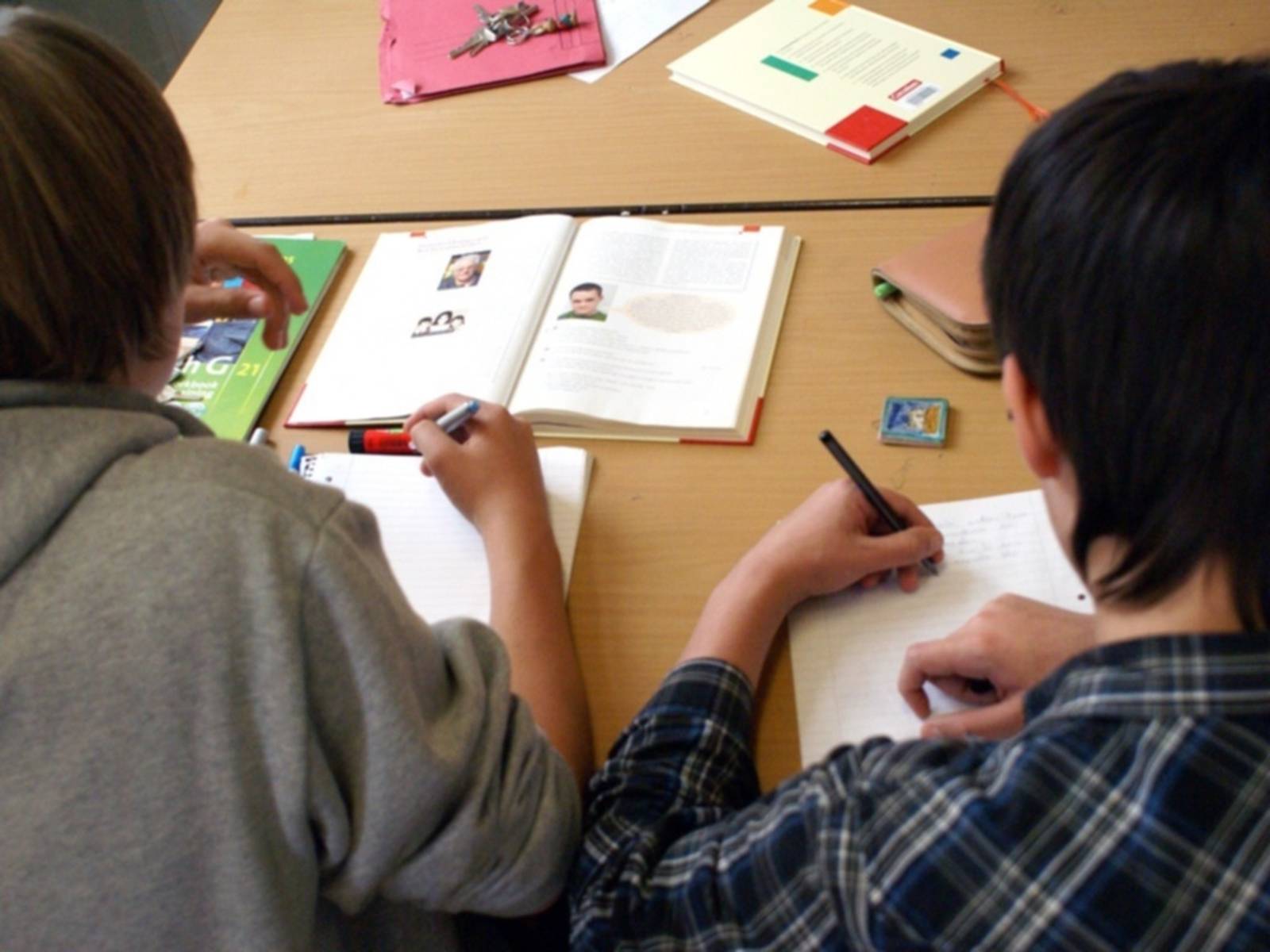 Über die Schultern von zwei Jugendlichen hinweg sind mehrere Schulbücher zu sehen, zudem schreiben die Jugendlichen gerade etwas in ihre Collegeblöcke