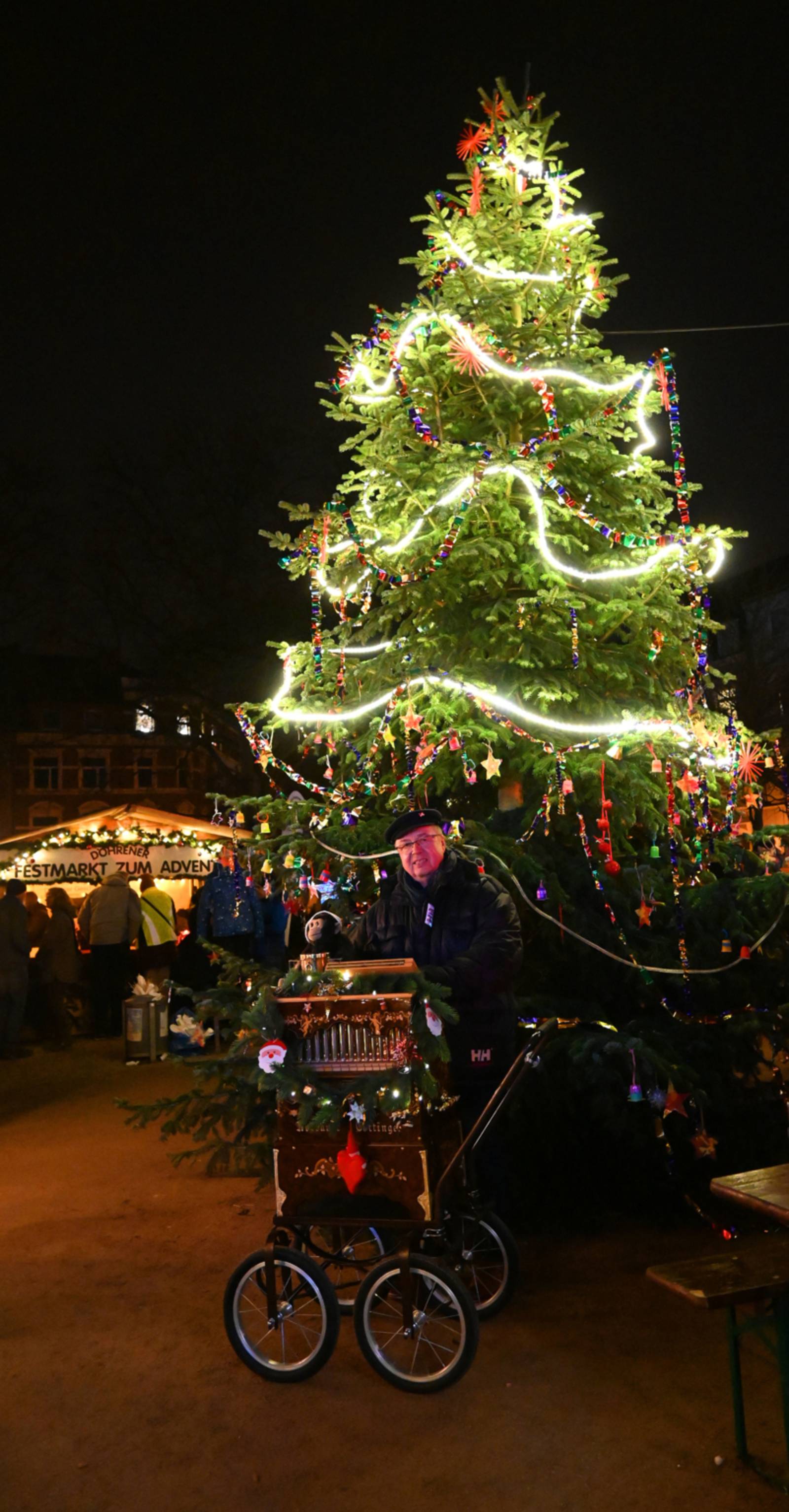 Auf dem Bild ist ein großer Weihnachtsbaum im Dunkeln zu sehen. Vor dem Weihnachtsbaum steht ein Straßen-Orgelspieler. Hinter dem Weihnachtsbaum ist eine Weihnachtsmarktbude mit der Aufschrift: "DÖHRENER FESTMARKT ZUM ADVENT" zu sehen.