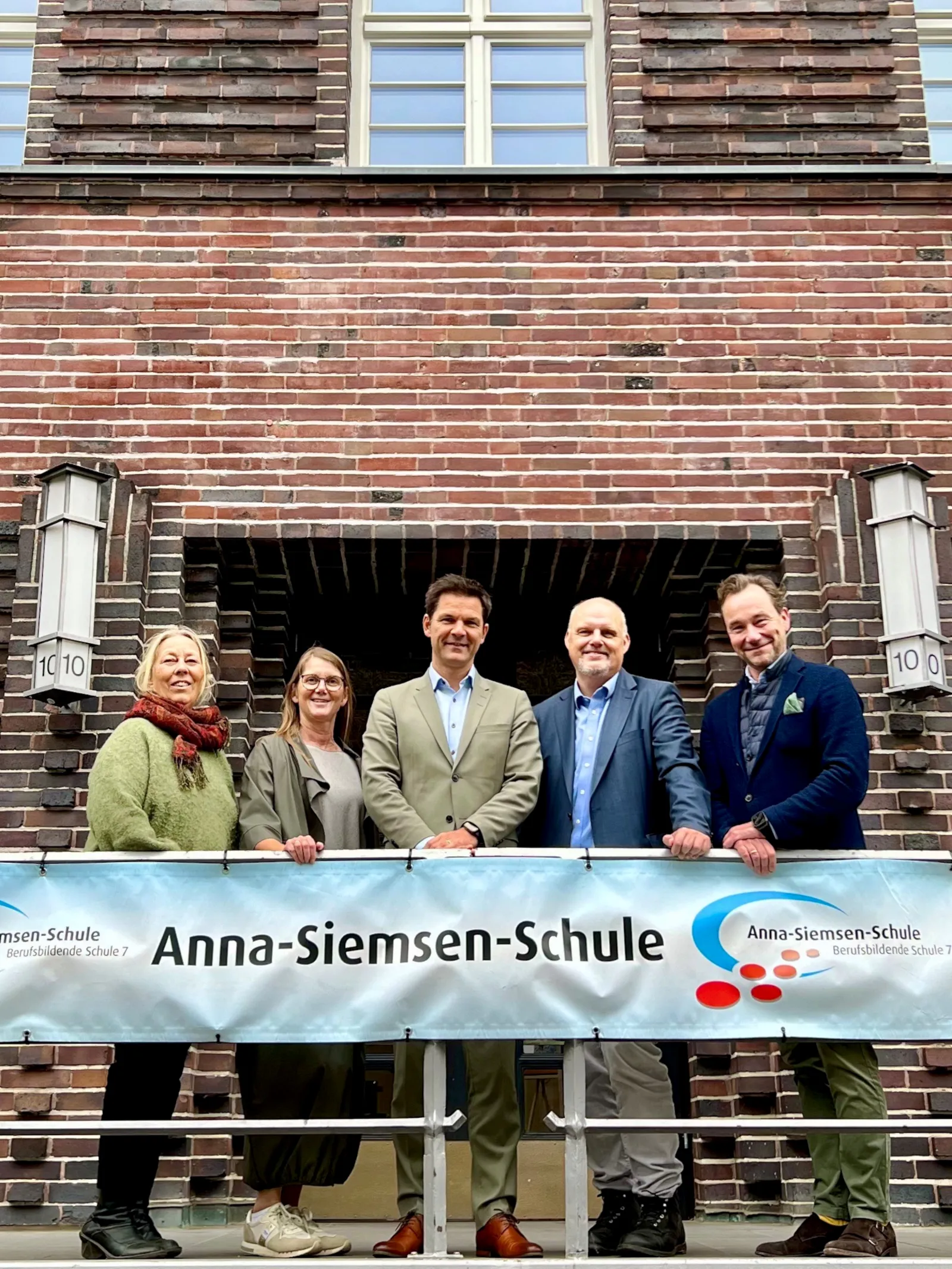 F&uuml;nf Personen stehen hinter einem Banner mit der Aufschrift "Anna-Siemsen-Schule" und vor dem Eingang eines Geb&auml;udes mit der Hausnummer 10.