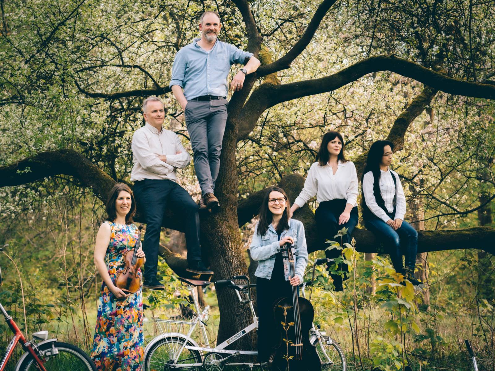 Vier Frauen und zwei Männer haben sich vor einem Baum und dessen Ästen verteilt. Im Bild sind außerdem Fahrräder und Musikinstrumente.