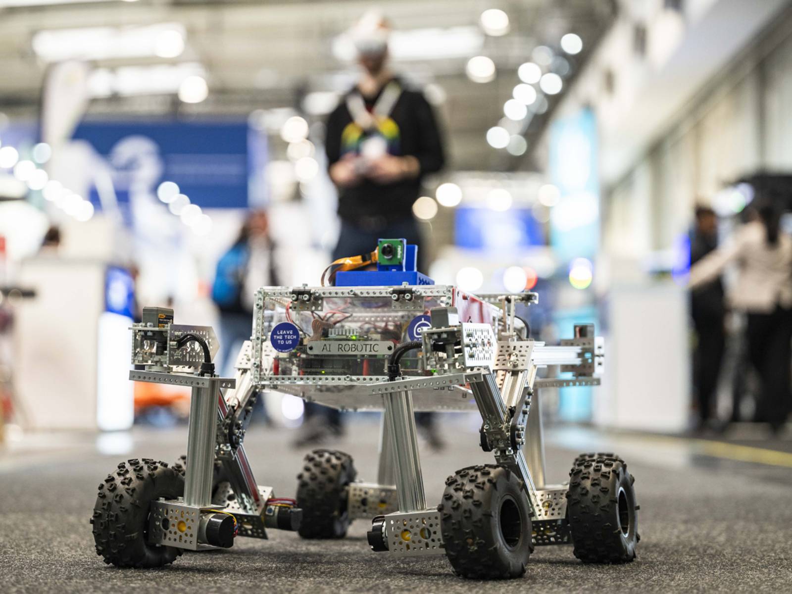Mann unscharf im Hintergrund mit VR-Brille und Fernsteuerung lenkt einen kleines metallenes Fahrzeug im Vordergrund.