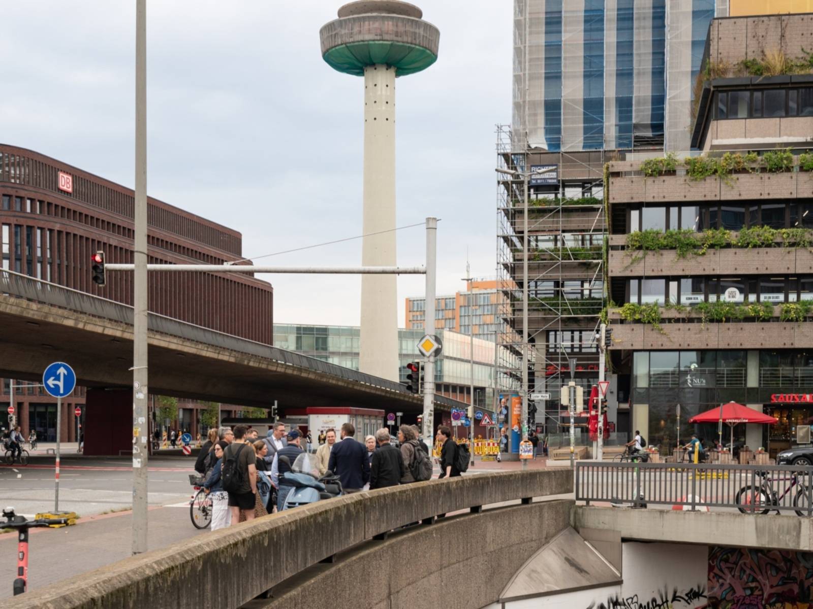 Menschen stehen auf einem Platz, links eine Kreuzung unter einer Hochstraße, hinten in der Mitte ein großer Funkturm, rechts hinten ein Gebäude, vorn rechts eine tiefere Untergeschossebene.