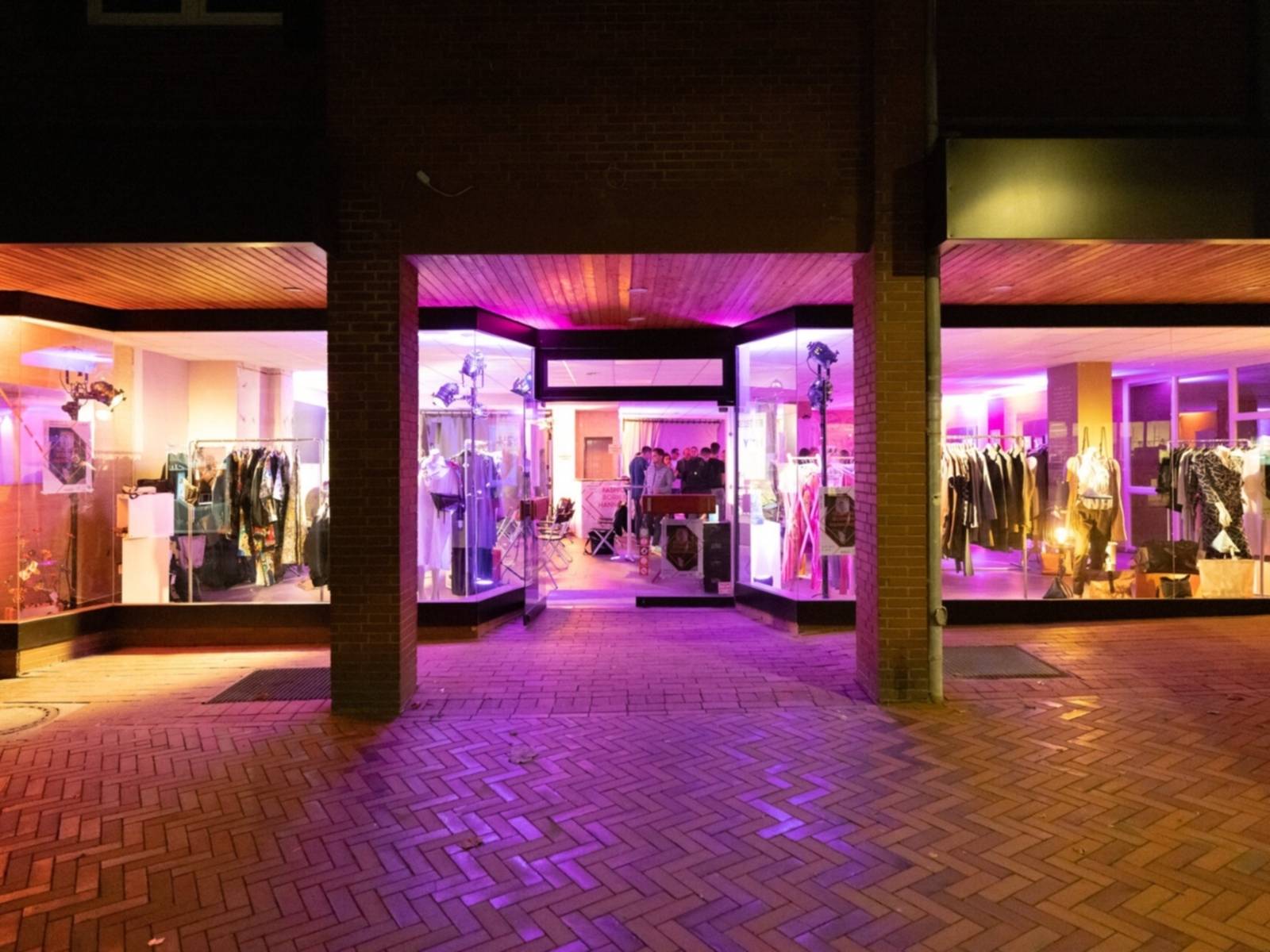 Eine Schaufensterzeile bei Nacht in lila Neonlicht. Im Ladengeschäft sind hauptsächlich Kleidungsstücke zu sehen.