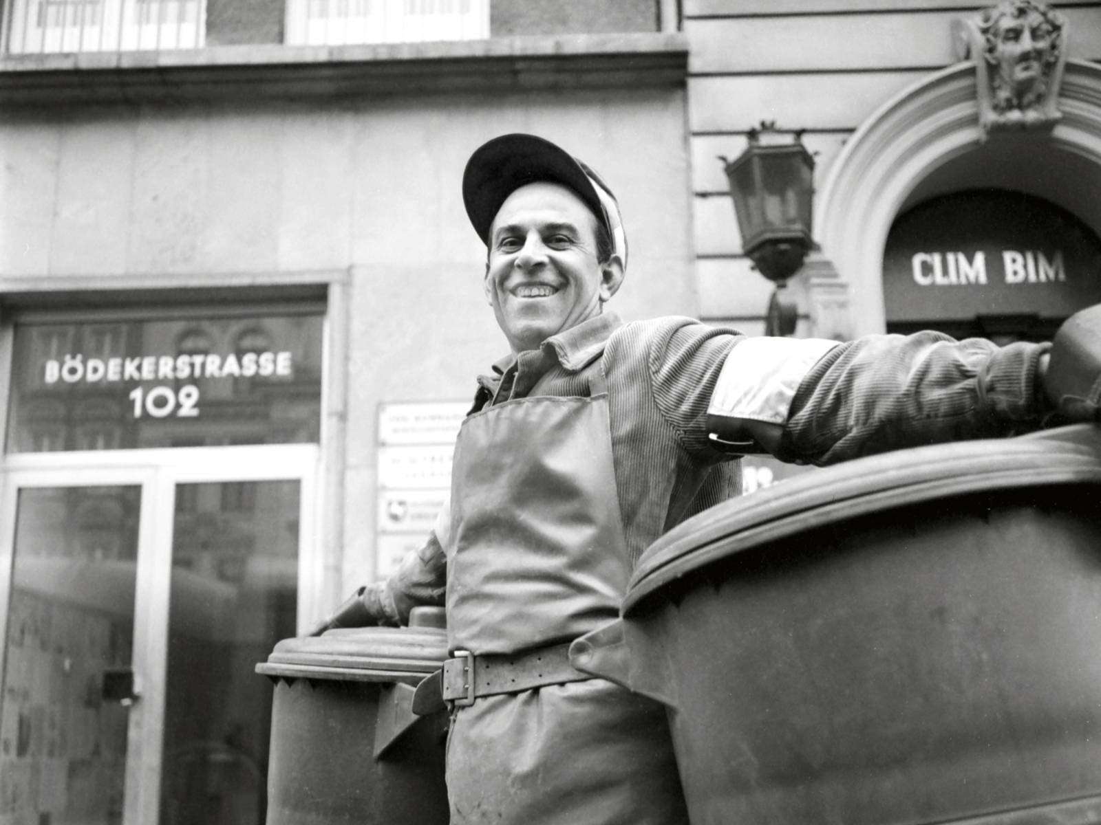Schwarz-Weiß-Fotografie: Ein Mann trägt eine grobe Schürze, unter jedem Arm hat er eine Mülltonne, die er drehend über die Straße bewegt.