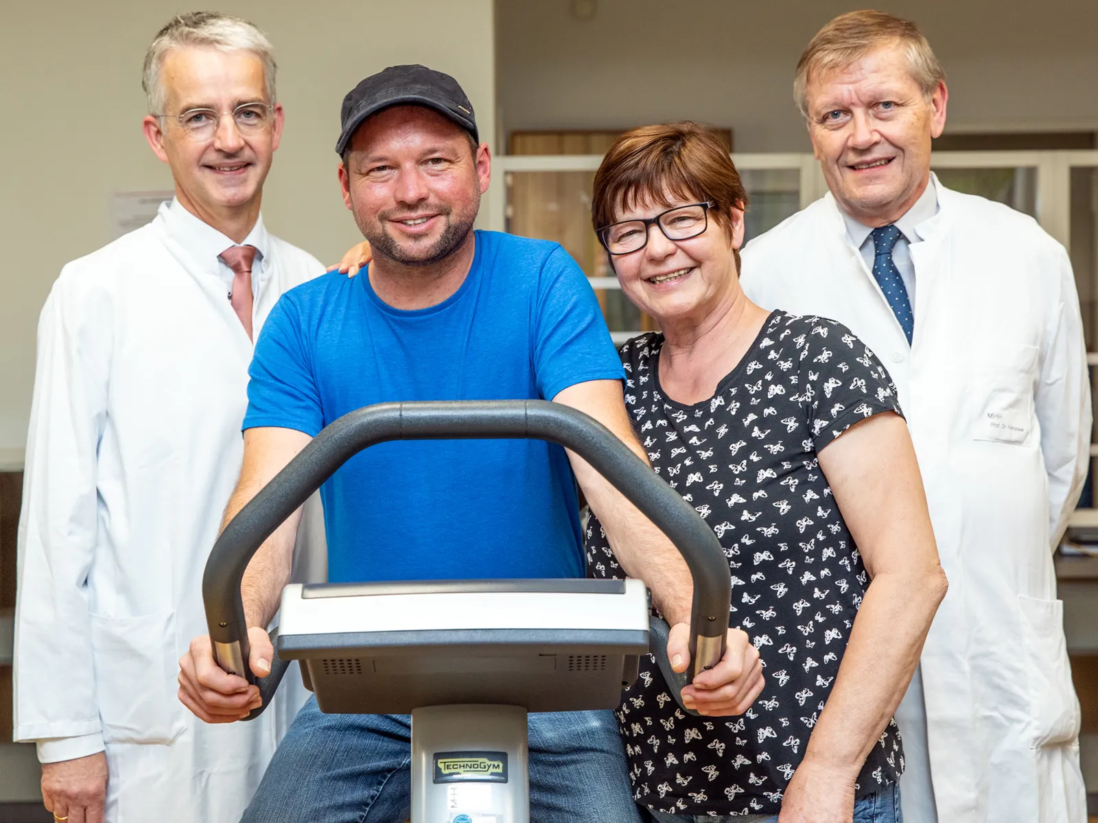 Mann auf einem Fitness-Fahrrad, daneben eine Frau und zwei Ärzte