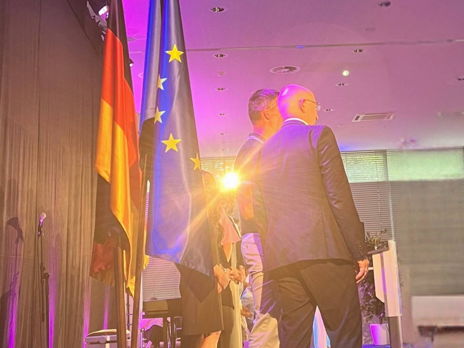 Links im Bild ist ein Flaggen-Ständer mit der europäischen, deutschen und Regionsflagge. Rechts davon ein Scheinwerfer der in die Kamera blendet, vor den Flaggen stehen mehrere Menschen.